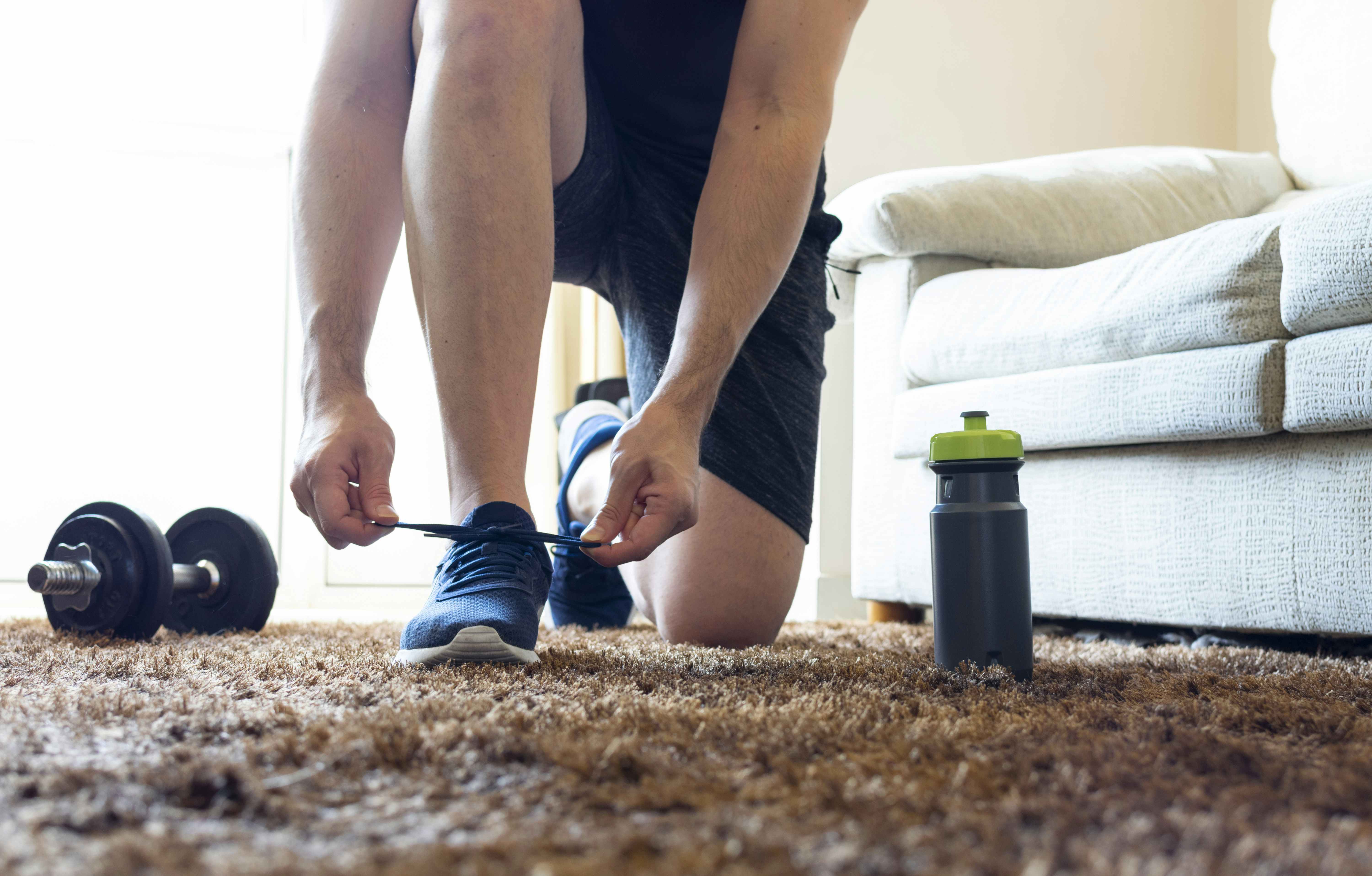A man tying his shoe on a rug at home next to weights and a water bottle