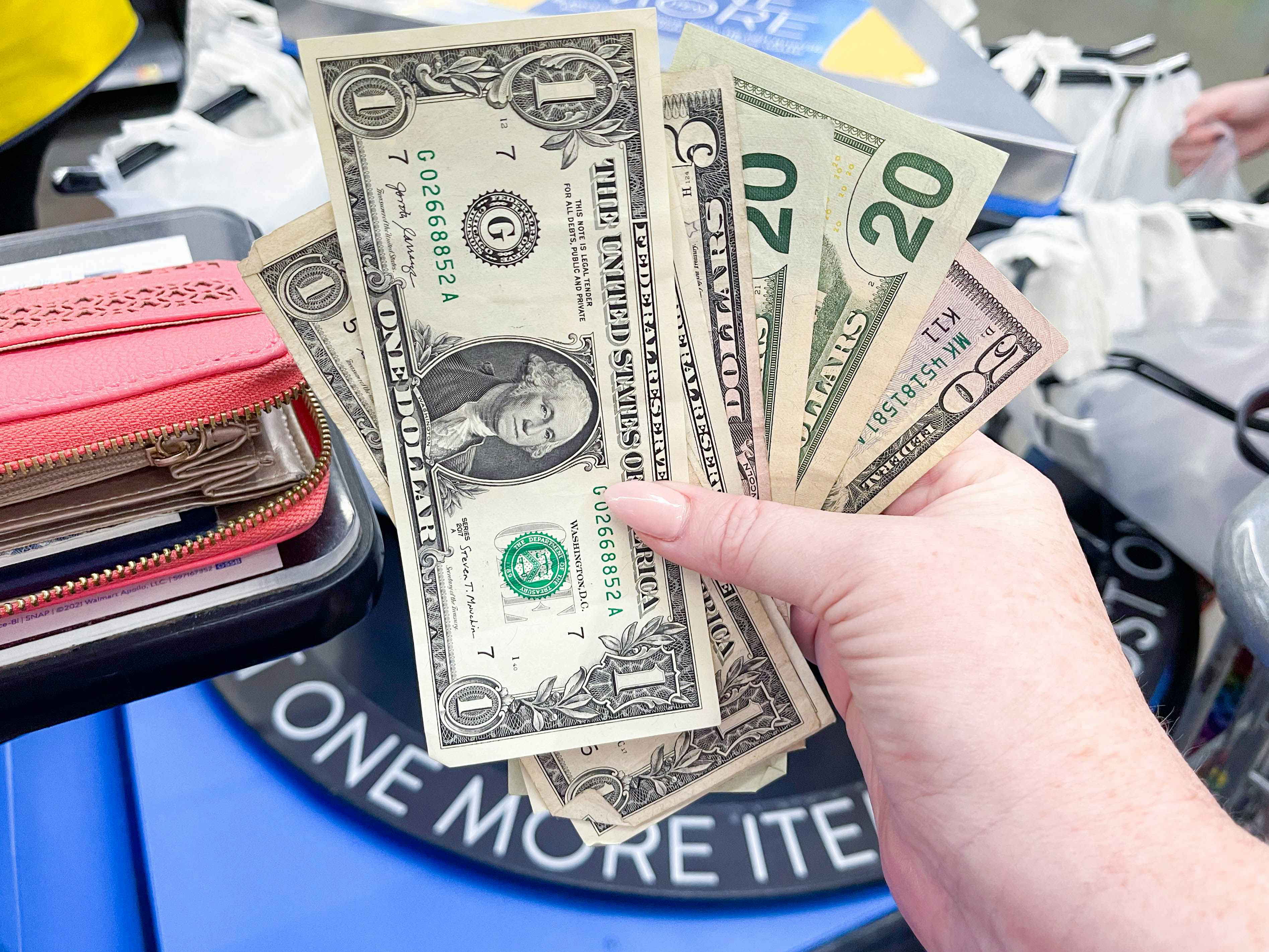 A person's hand holding up $98 in cash at the checkout lane in Walmart.