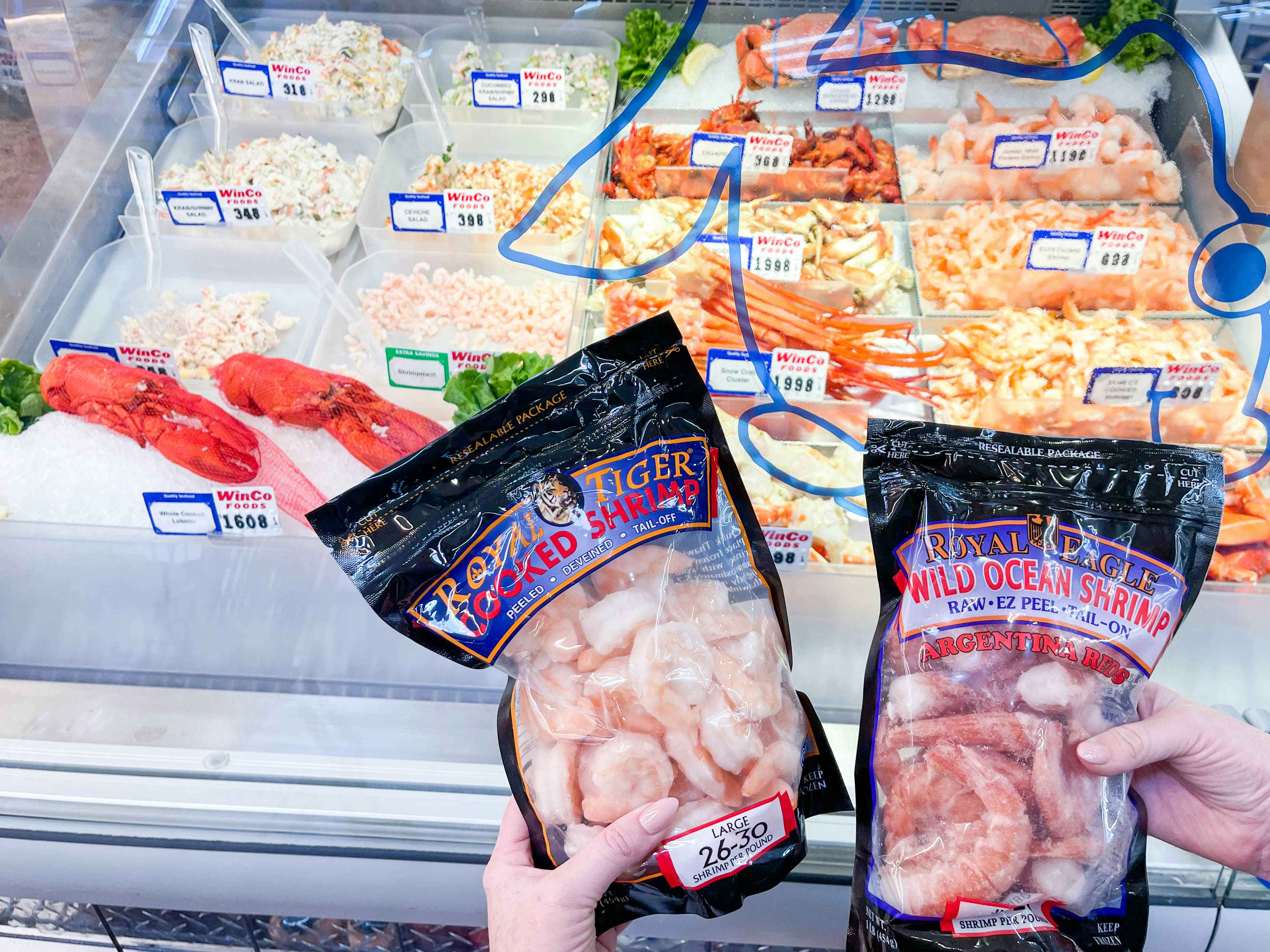 A person's hands holding up two bags of Royal Tiger Wild Ocean Shrimp in front of the seafood display at WinCo Foods.