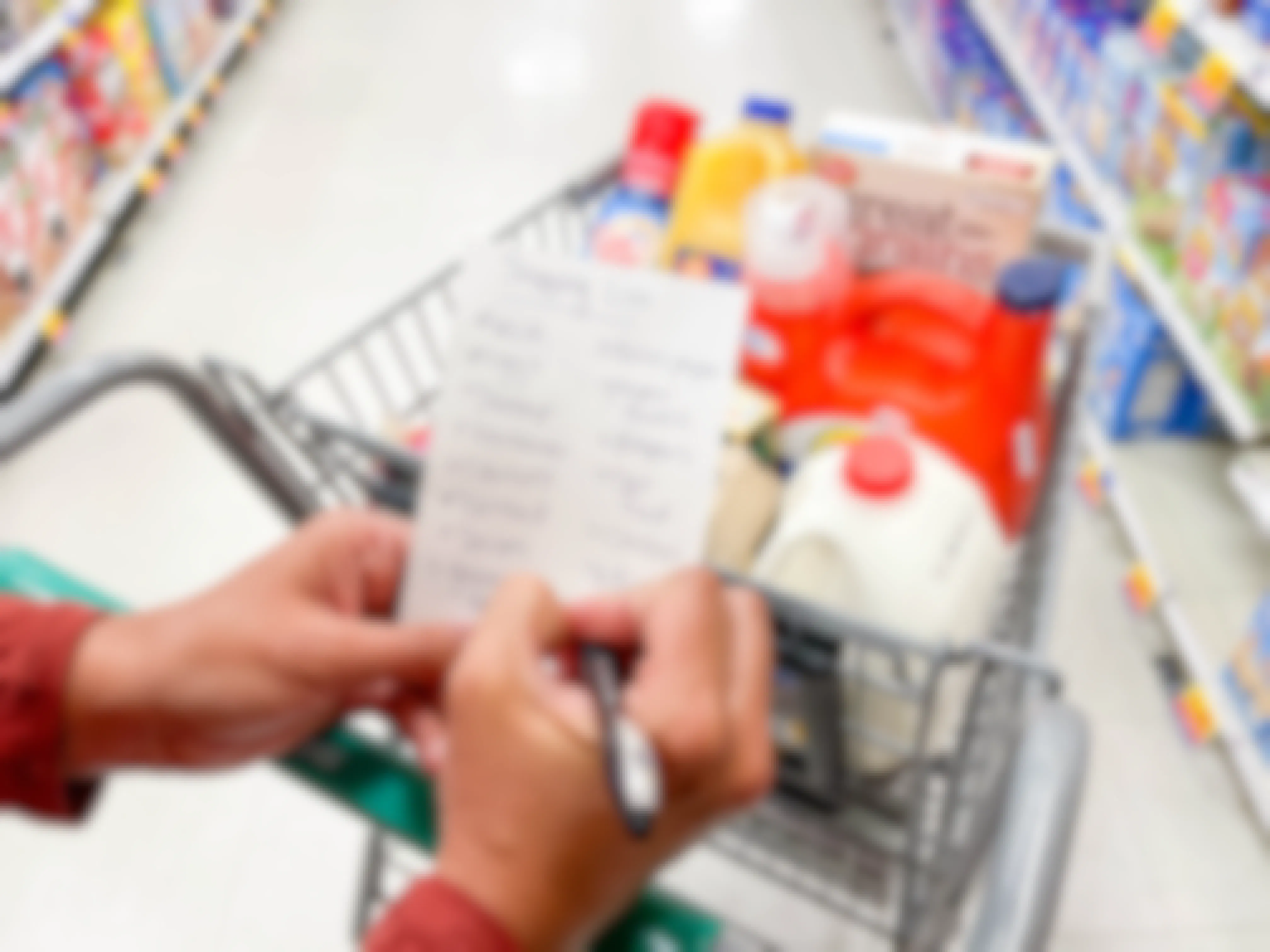 A woman at the grocery store, using a pen to check off an item on her shopping list while resting her hands and wrists on her shopping cart filled with milk, laundry detergent, orange juice, cereal, and other grocery items.