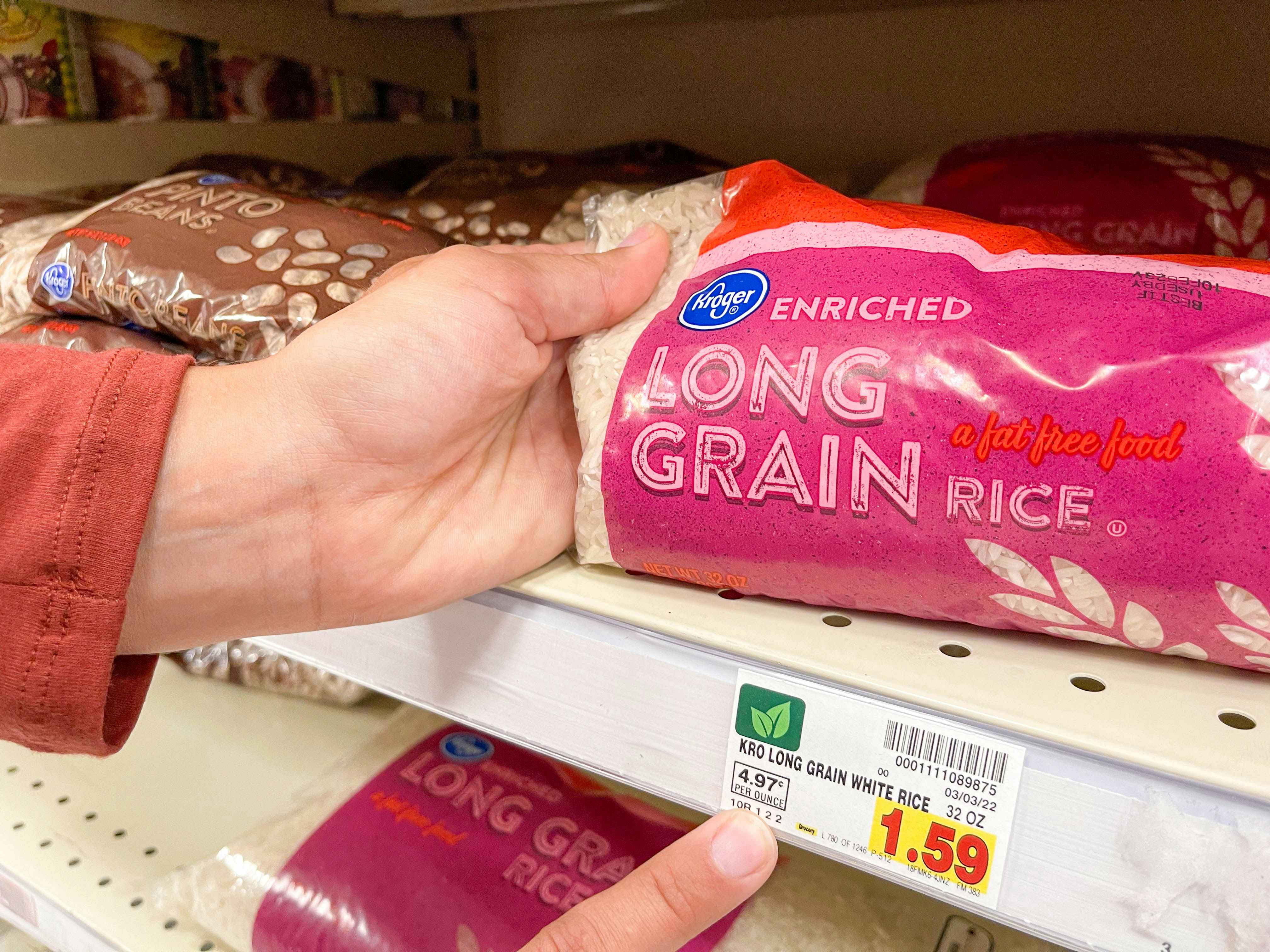 A person's hands, one holding up one end of a bag of Kroger enriched long grain rice sitting on a shelf, and the other hand pointing at the price tag for the displayed product.