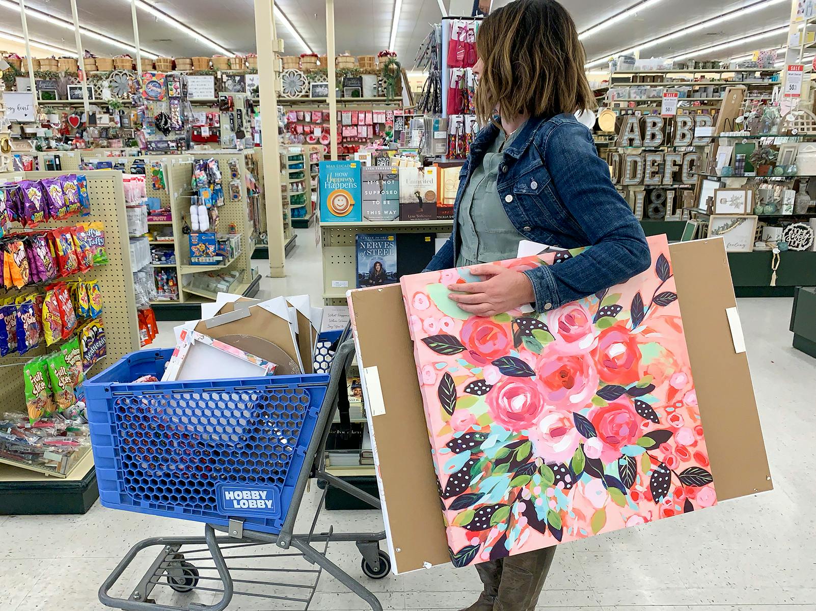 A woman standing the the checkout line with a cart full of products, holding two large art prints.