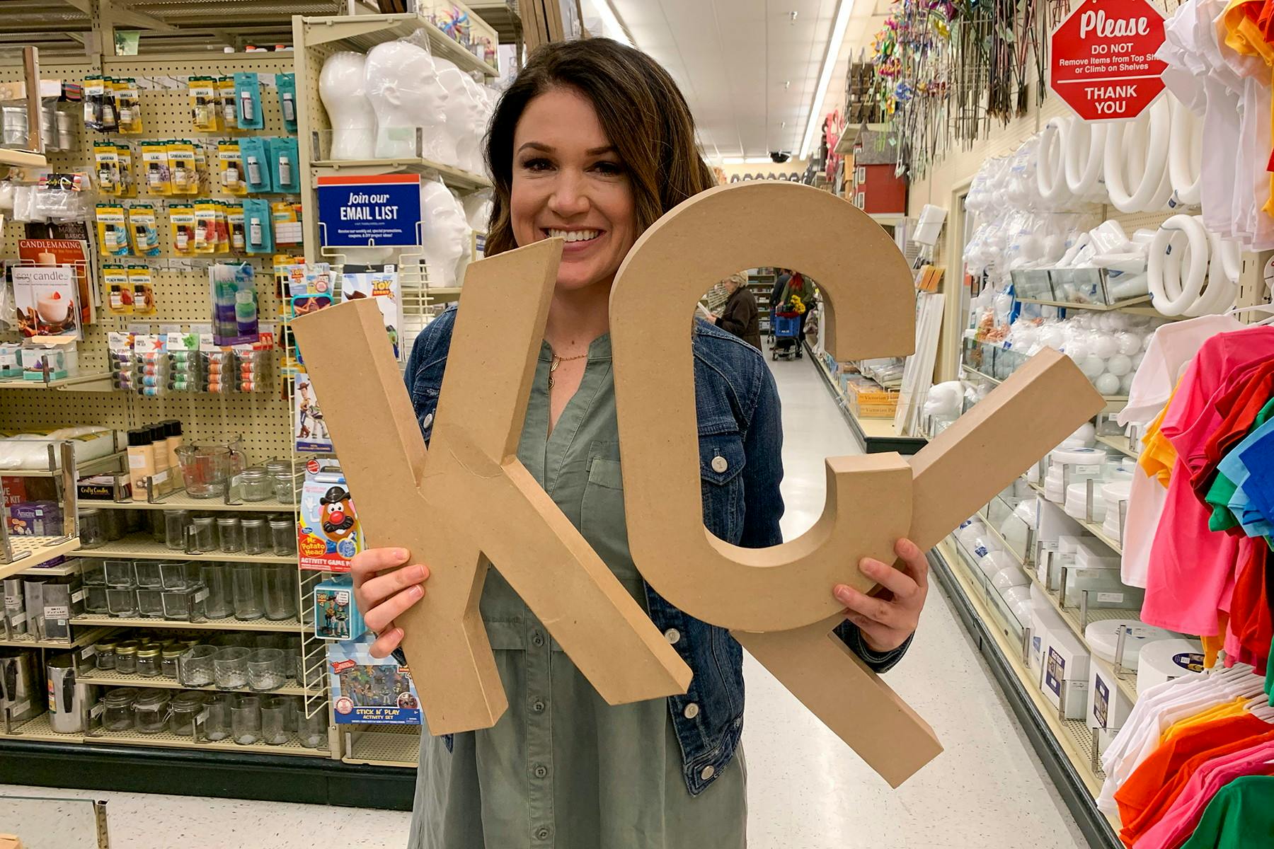 One of the owners of thekrazycouponlady.com, holding up large cardboard letters that read "KCL" in a craft store