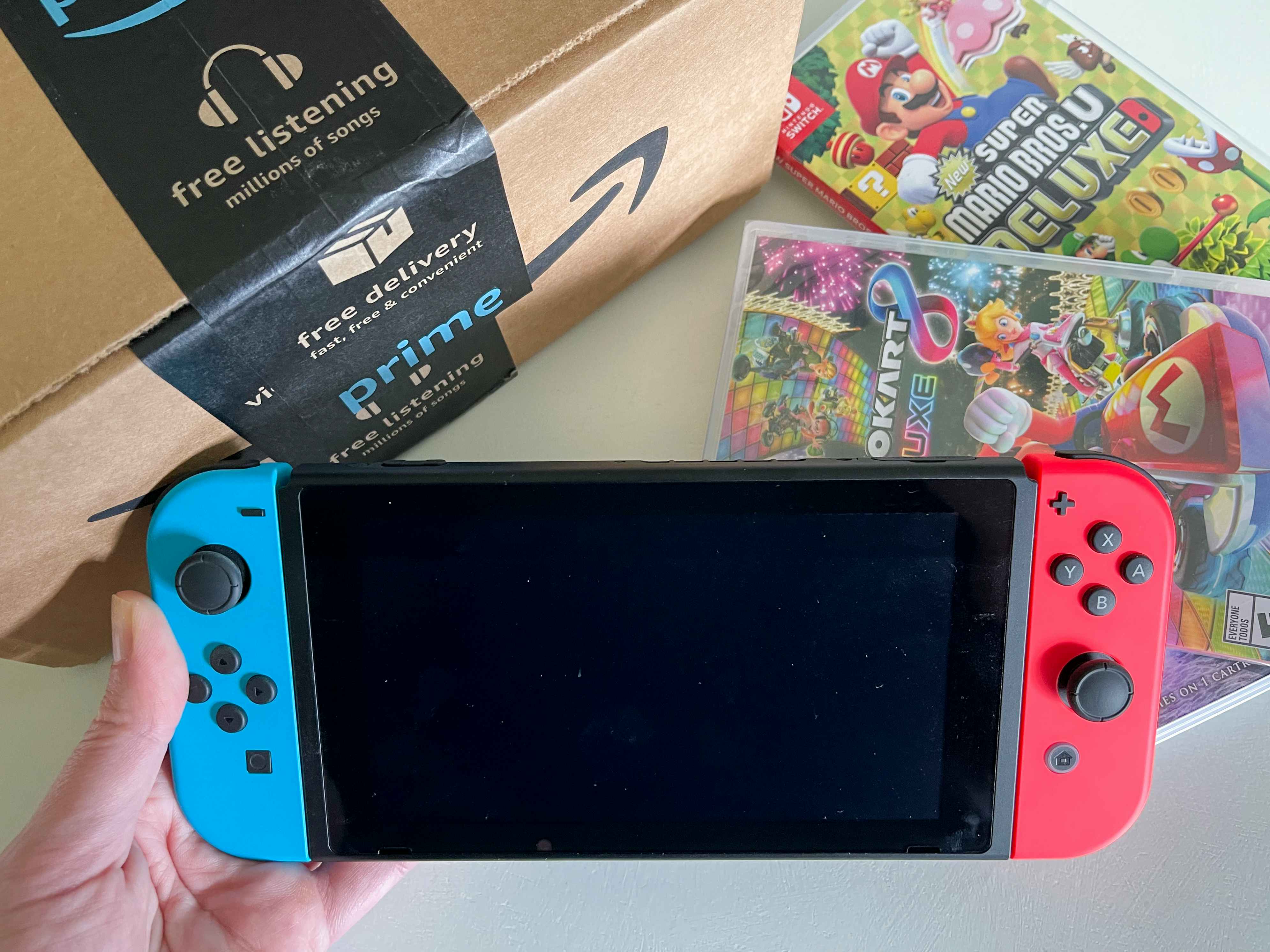 A person's hand holding a Nintendo Switch console next to an Amazon box and two Mario Switch games.