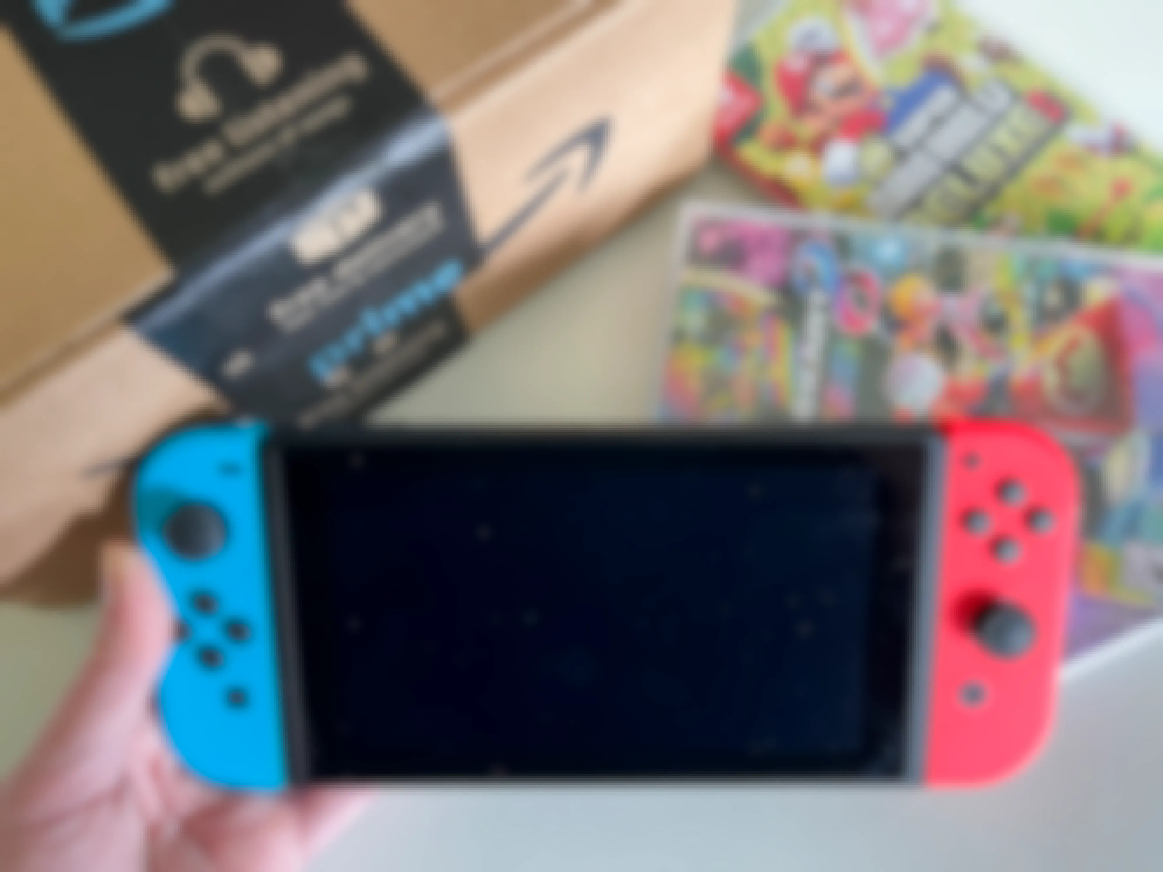A person's hand holding a Nintendo Switch console next to an Amazon box and two Mario Switch games.