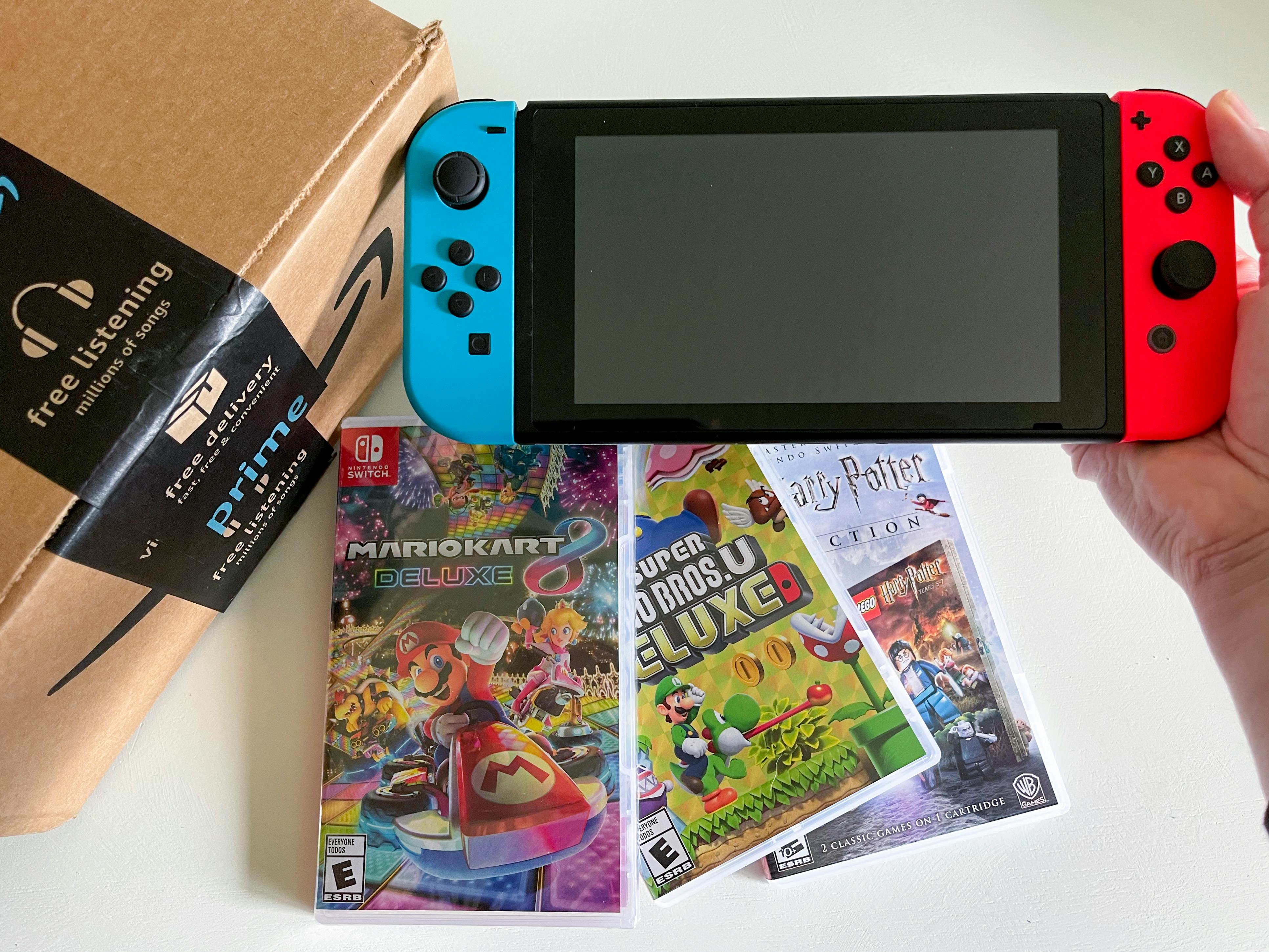 A person's hand holding up a Nintendo Switch next to an Amazon box with three Switch games below it.