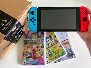 A person's hand holding up a Nintendo Switch next to an Amazon box with three Switch games below it.