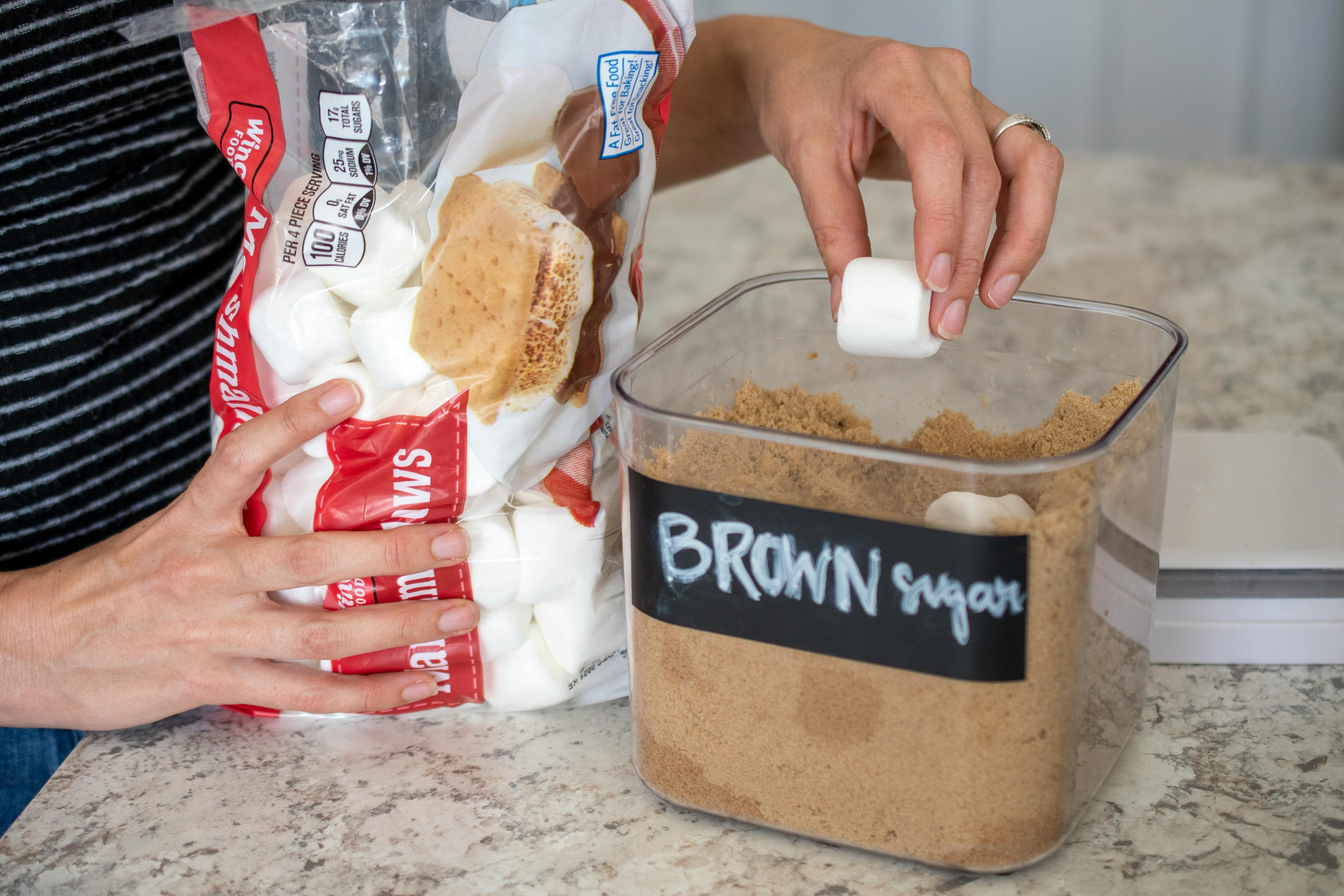 A person putting two large marshmallows inside a container filled with brown sugar.