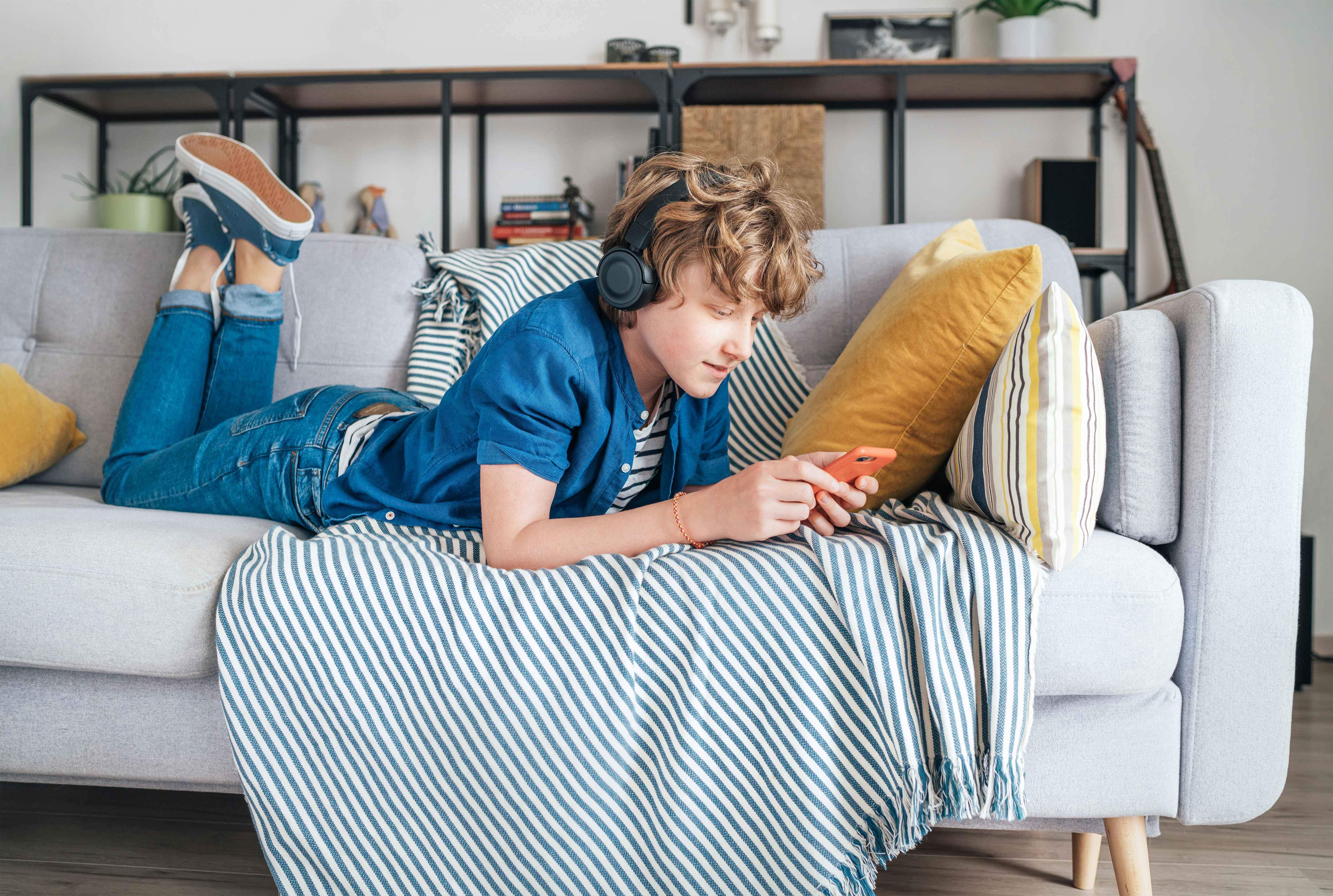 Preteen boy lying at home on cozy sofa dressed casual jeans and shirt. listening to music and chatting using wireless headphones connected with smartphone. Child using electronic devices concept