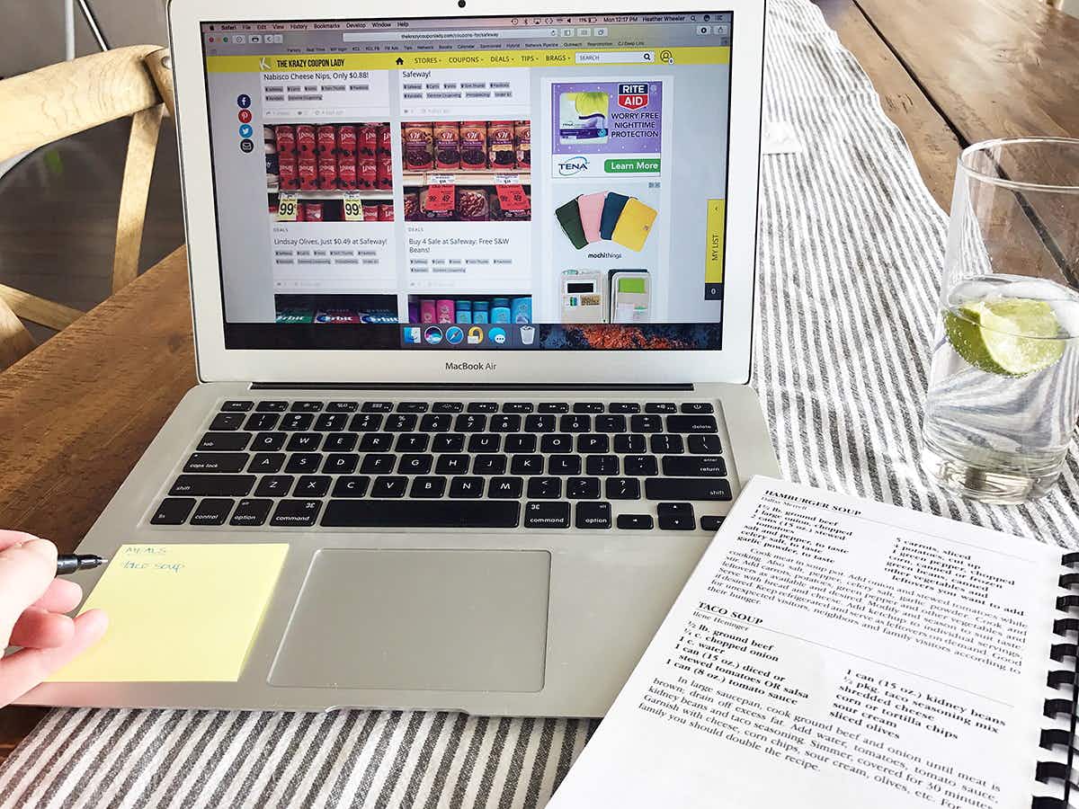 Closeup of a laptop opened to KCL deals. There's a printed paper with recipes and a hand making a list on a sticky note.