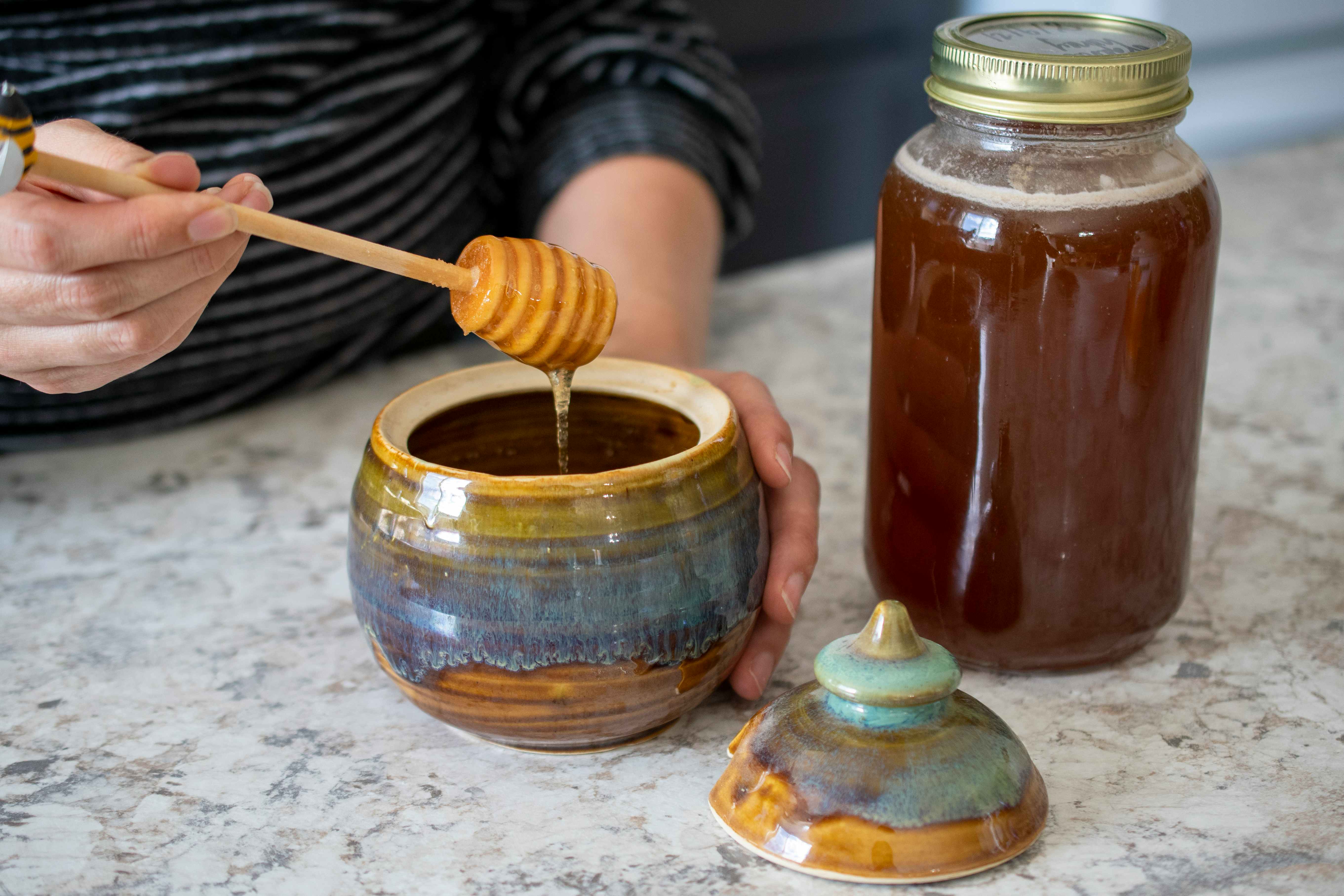A person scooping honey from a honey pot with a honey stir stick 