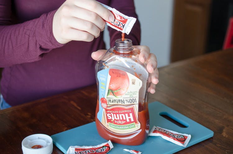 A person dumping Ketchup packets into a Hunts Tomato Ketchup bottle.