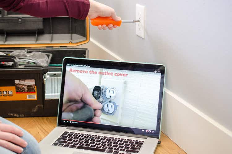 Person following instruction on youtube video on computer, removing outlet cover with a screw driver.