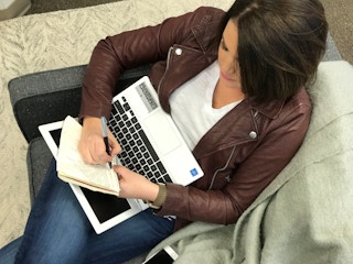 woman sitting on a couch on her laptop and writing on a notebook