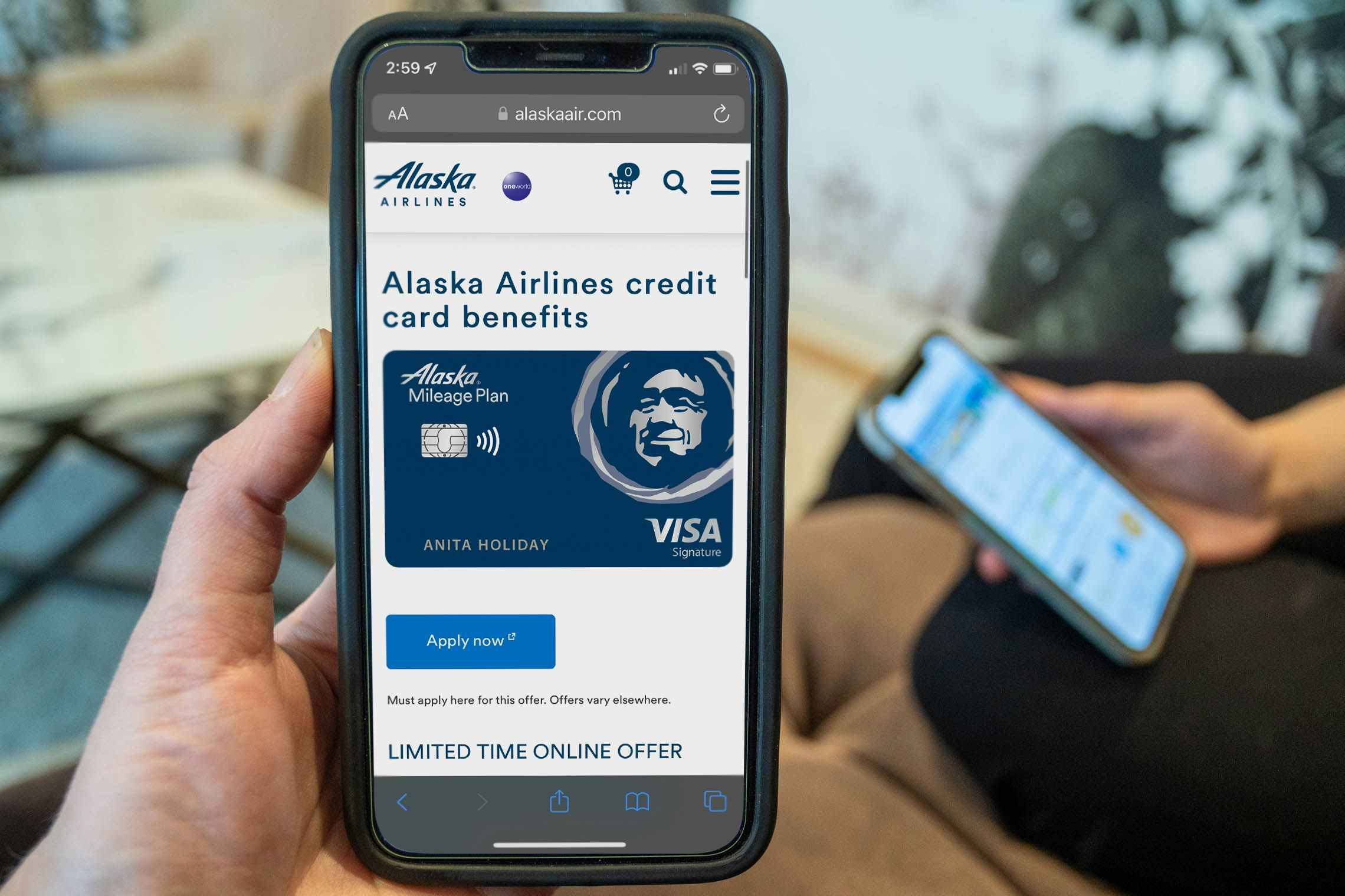 A person holding a phone with the Alaska Airlines visa card information displayed with someone else holding their own phone in the background