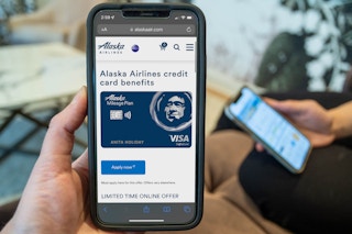 A person holding a phone with the Alaska Airlines visa card information displayed with someone else holding their own phone in the background