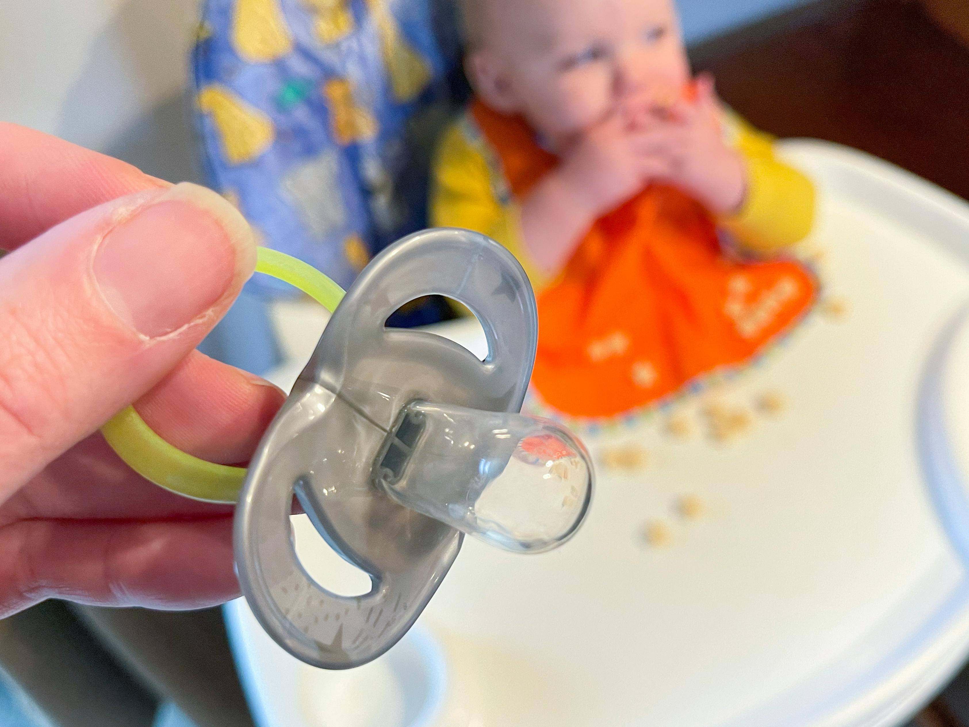 A pacifier in a persons hand with a baby in the background