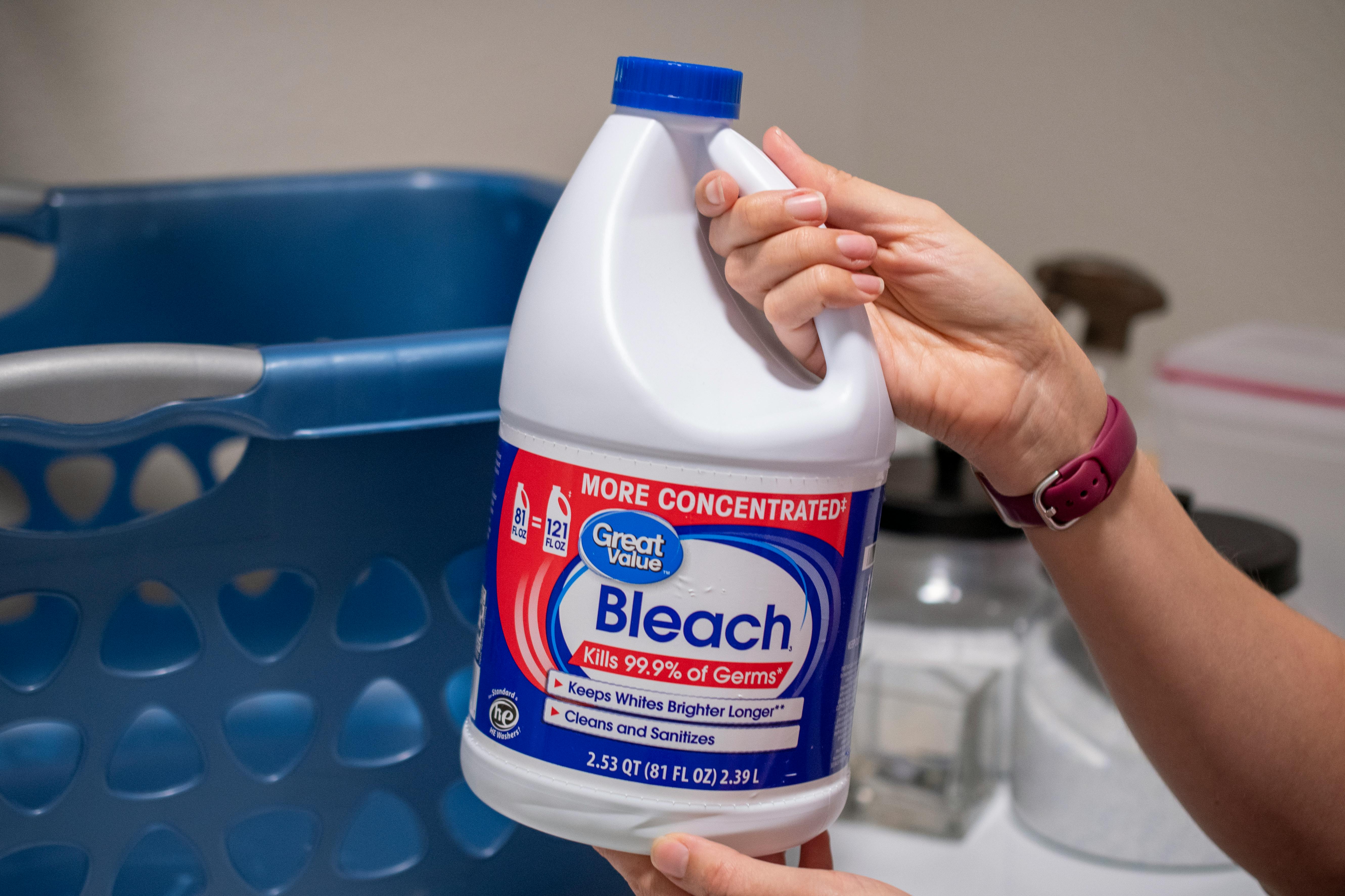 A person holding a bottle of bleach next to a washing machine.