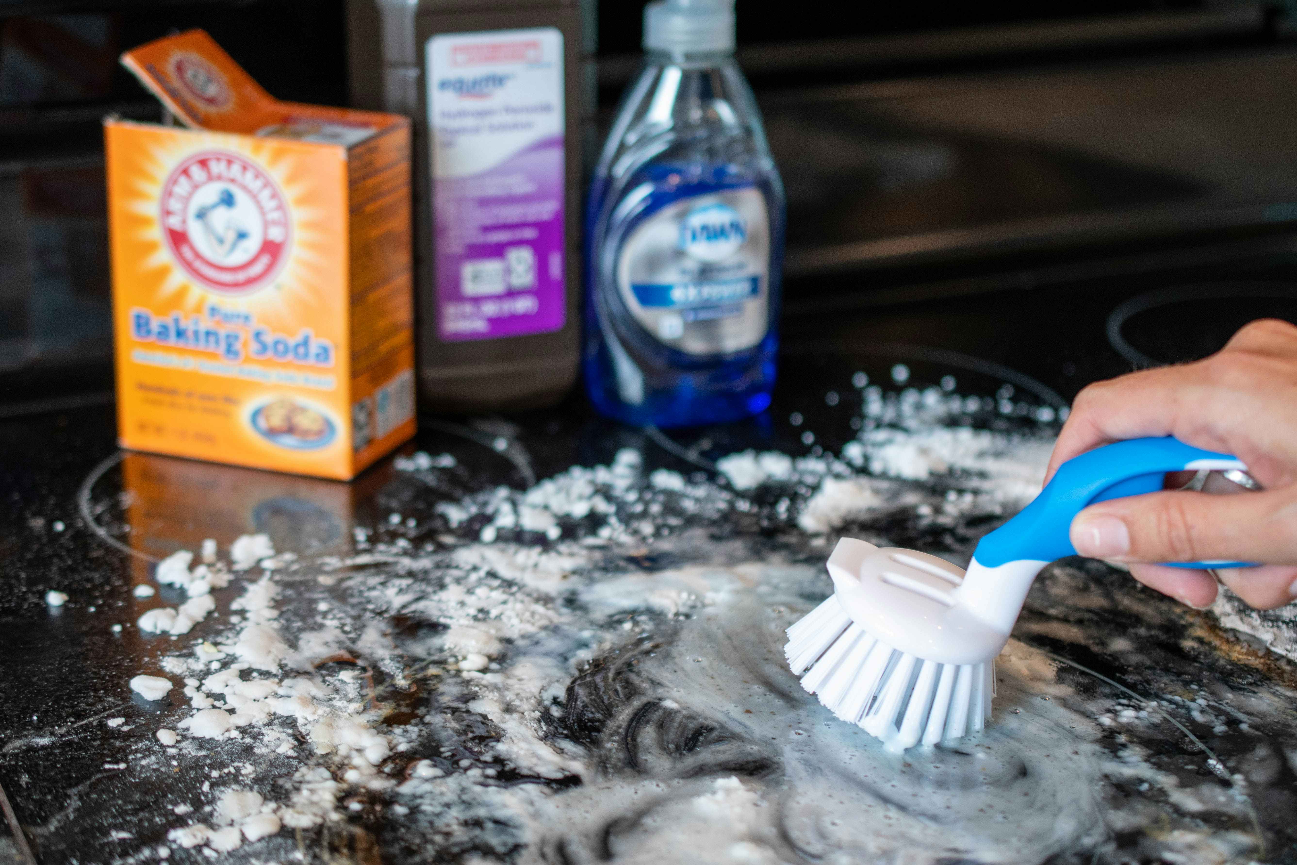 A person scrubbing a glass stove top with making soda, hydrogen peroxide, and dawn dish soap