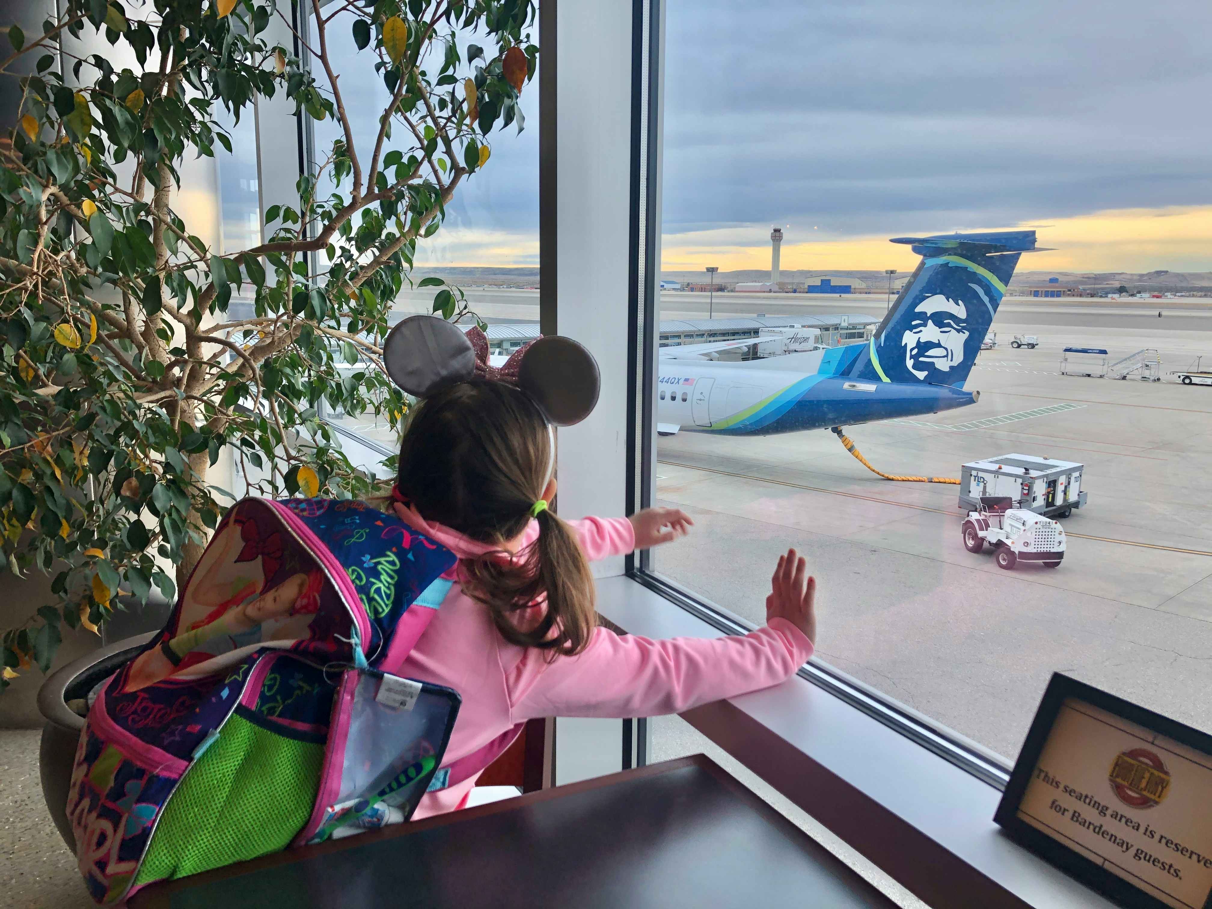 A little girl with Minnie Mouse ears looks out the window at the airport at Alaskan airlines.
