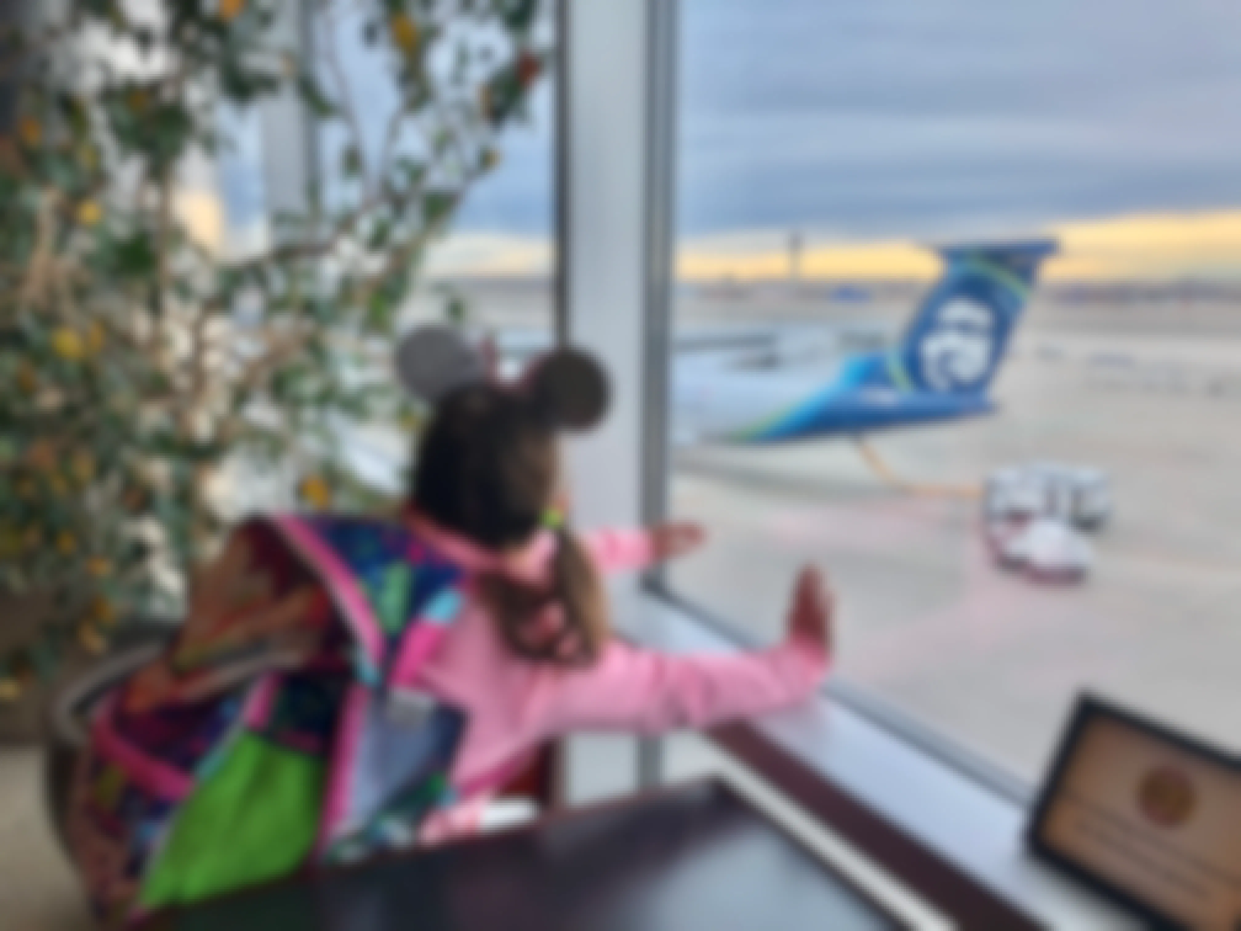 A little girl with Minnie Mouse ears looks out the window at the airport at Alaskan airlines plane.