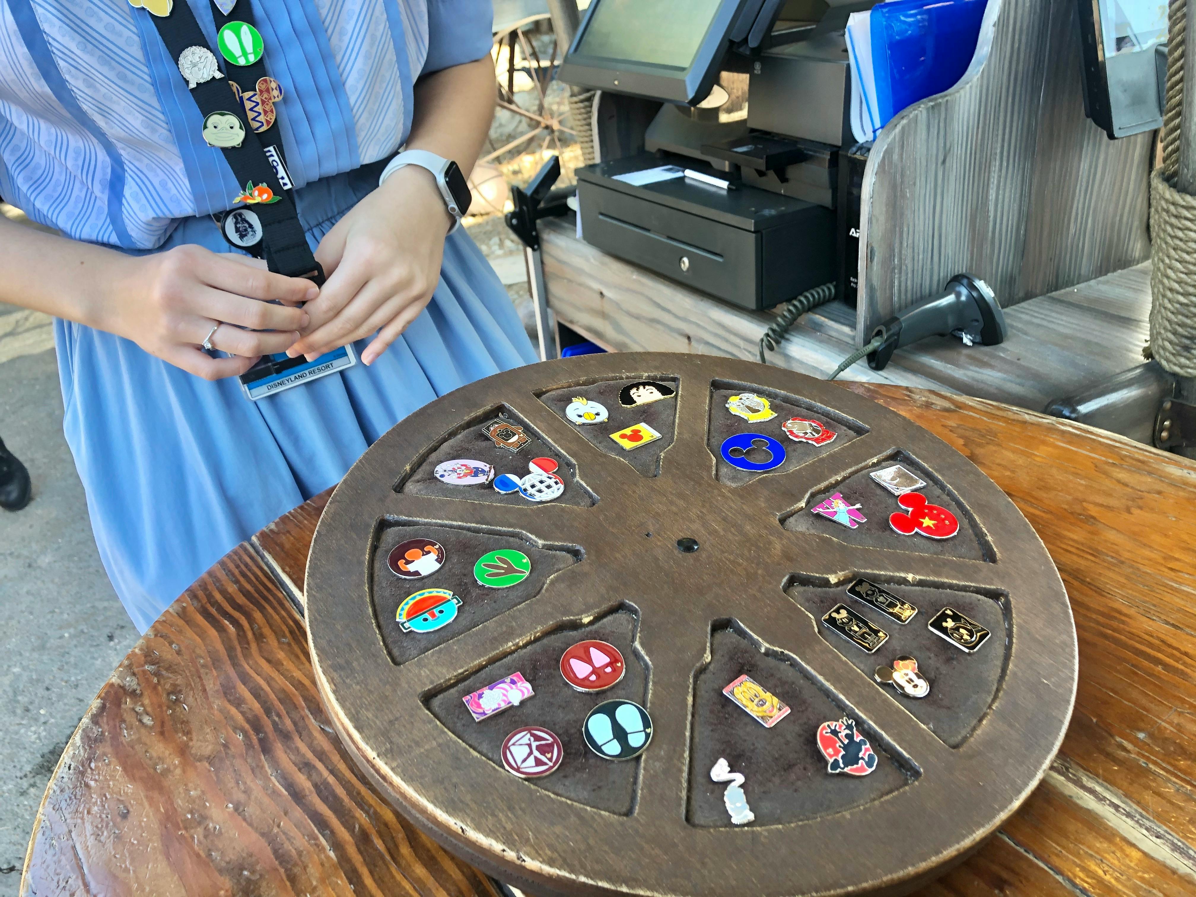 A person with a lanyard of pins standing near a wheel with pins on it at a Disney park.