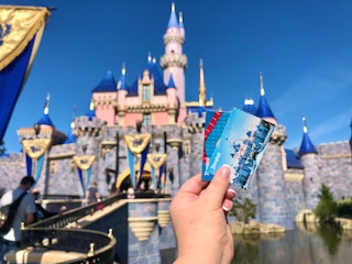 Disneyland tickets are held up in front of the Disneyland castle.