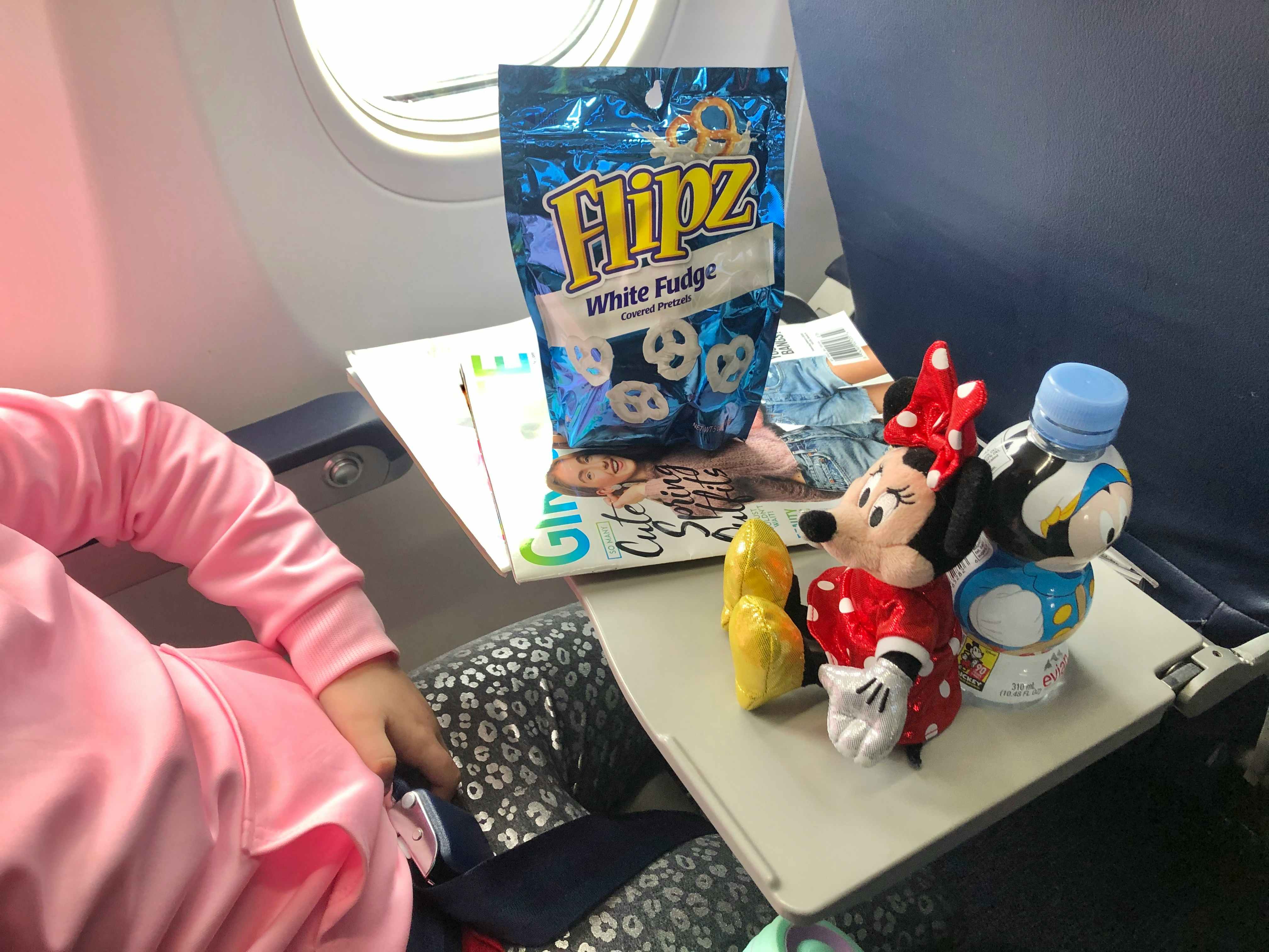 A girl sits in an airplane with a Mini mouse doll and snacks on her tray table.