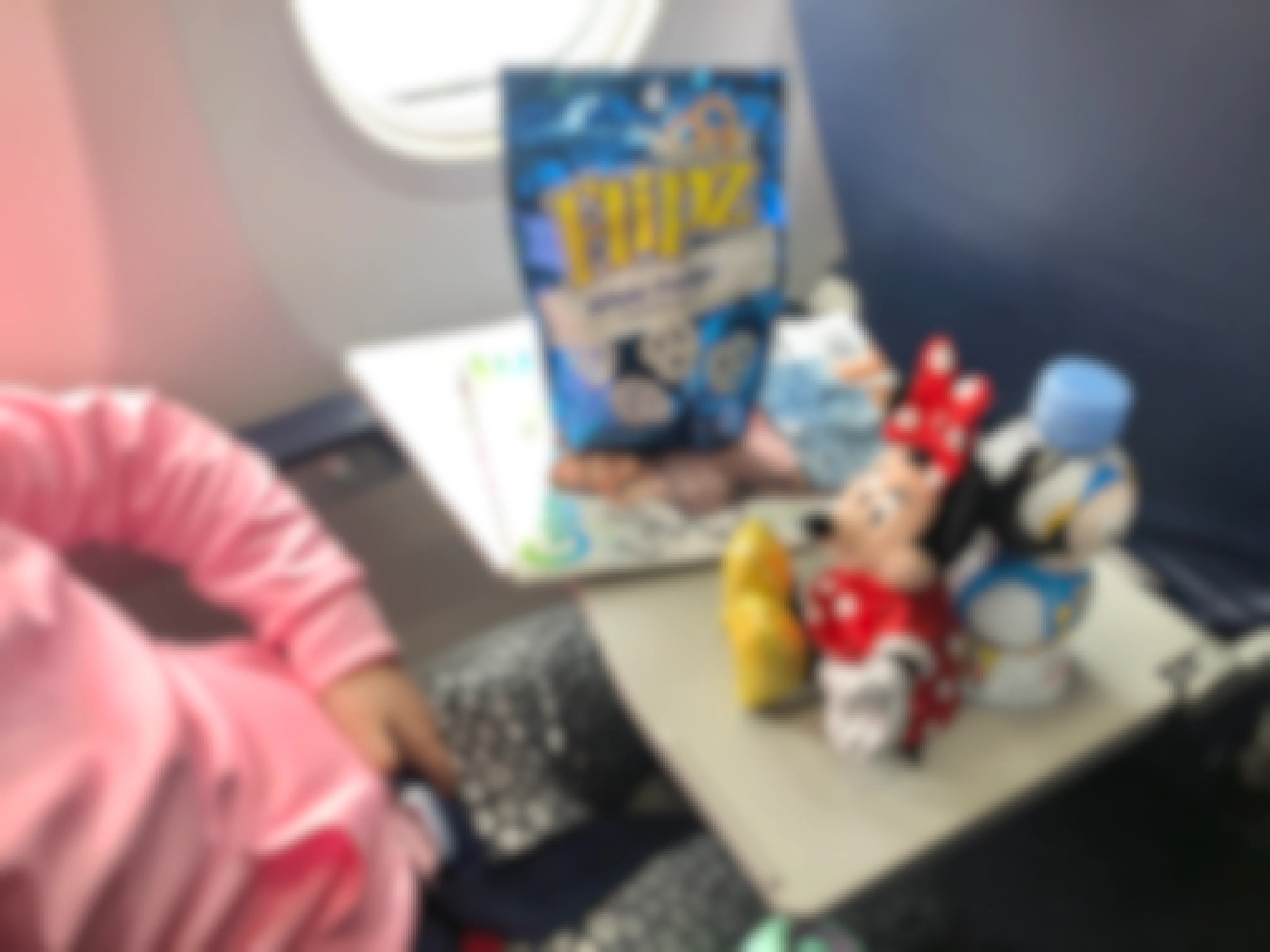 A girl sits in an airplane with a Mini mouse doll and snacks on her tray table.