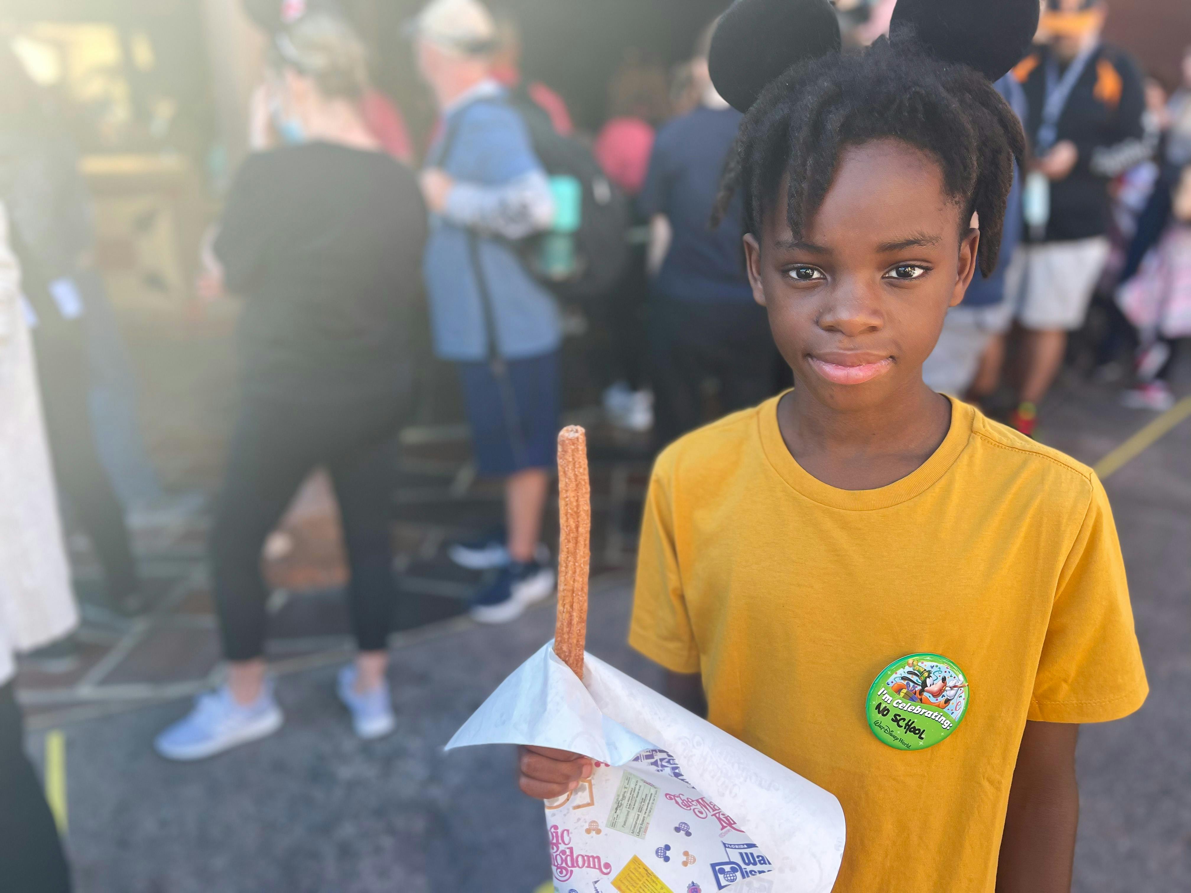 A child with Mickey Mouse ears and a celebration button standing in a line holding a churro.