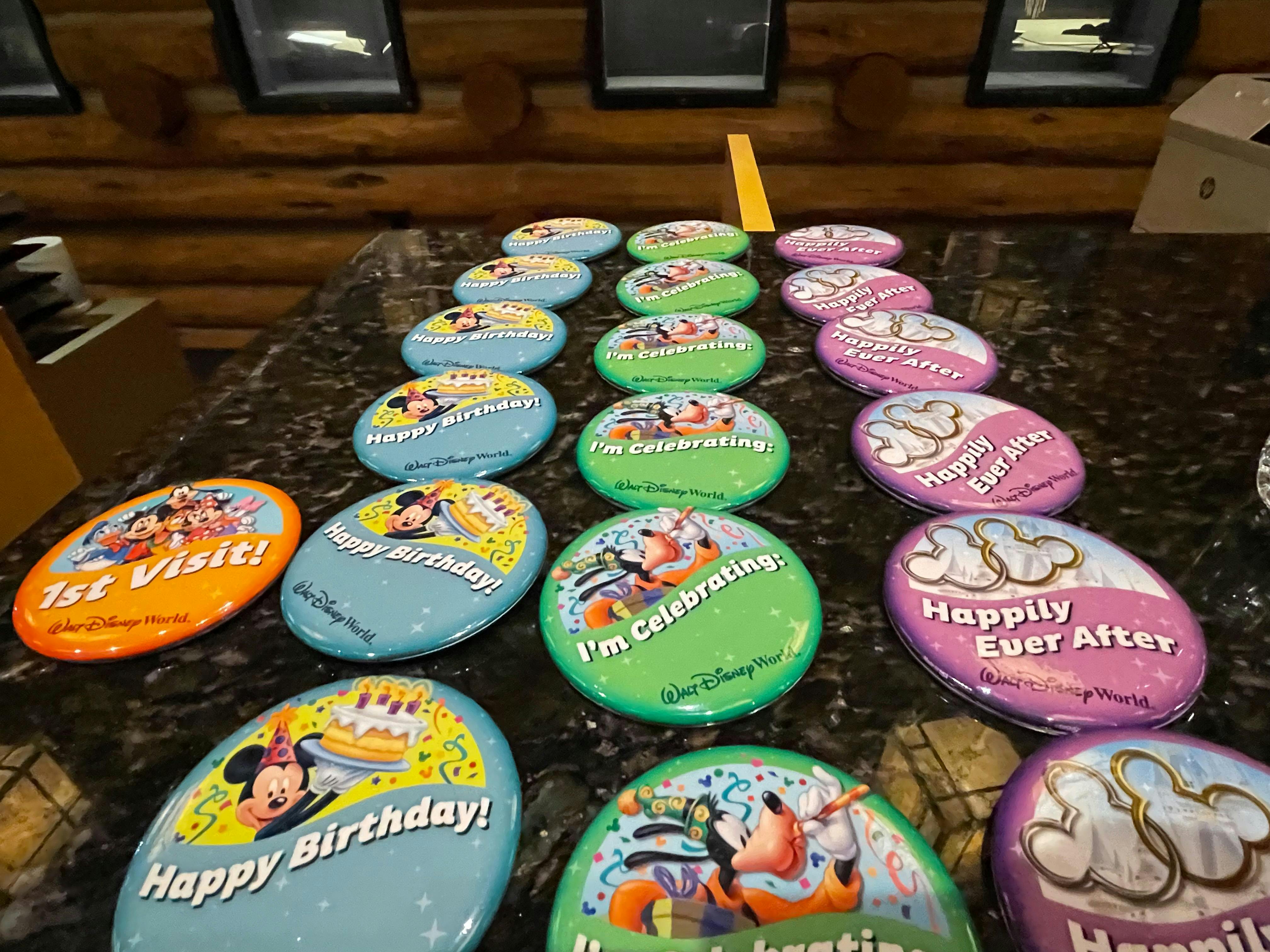Several different kinds of Disney celebration buttons sitting on a counter, including 1st Visit, Happy Birthday, I'm Celebrating, and Happily Ever After.