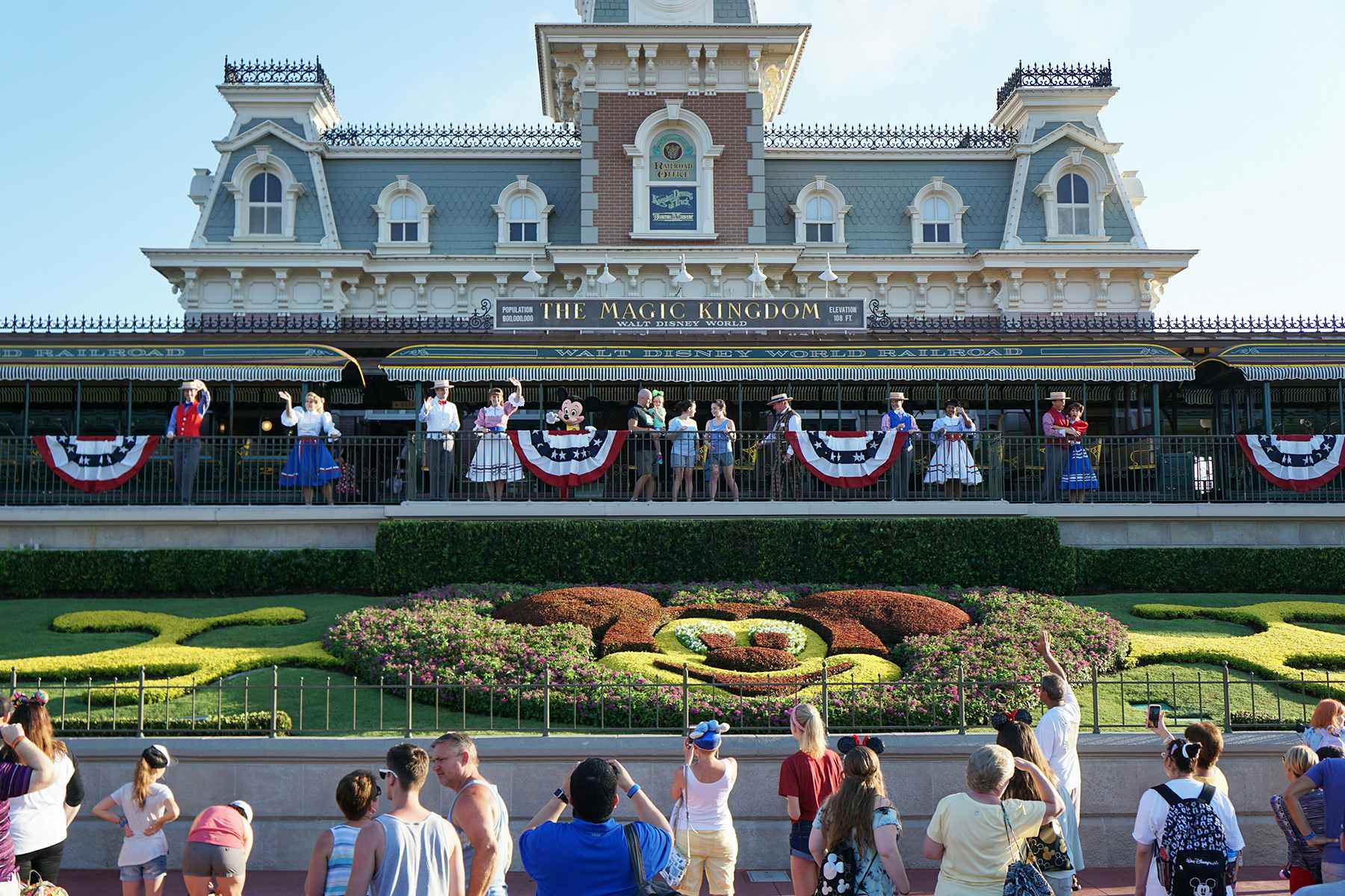 The train station building with America flag buntings and the Mickey Mouse head in the flower bed at the front of the Disney Magic Kingdom park.
