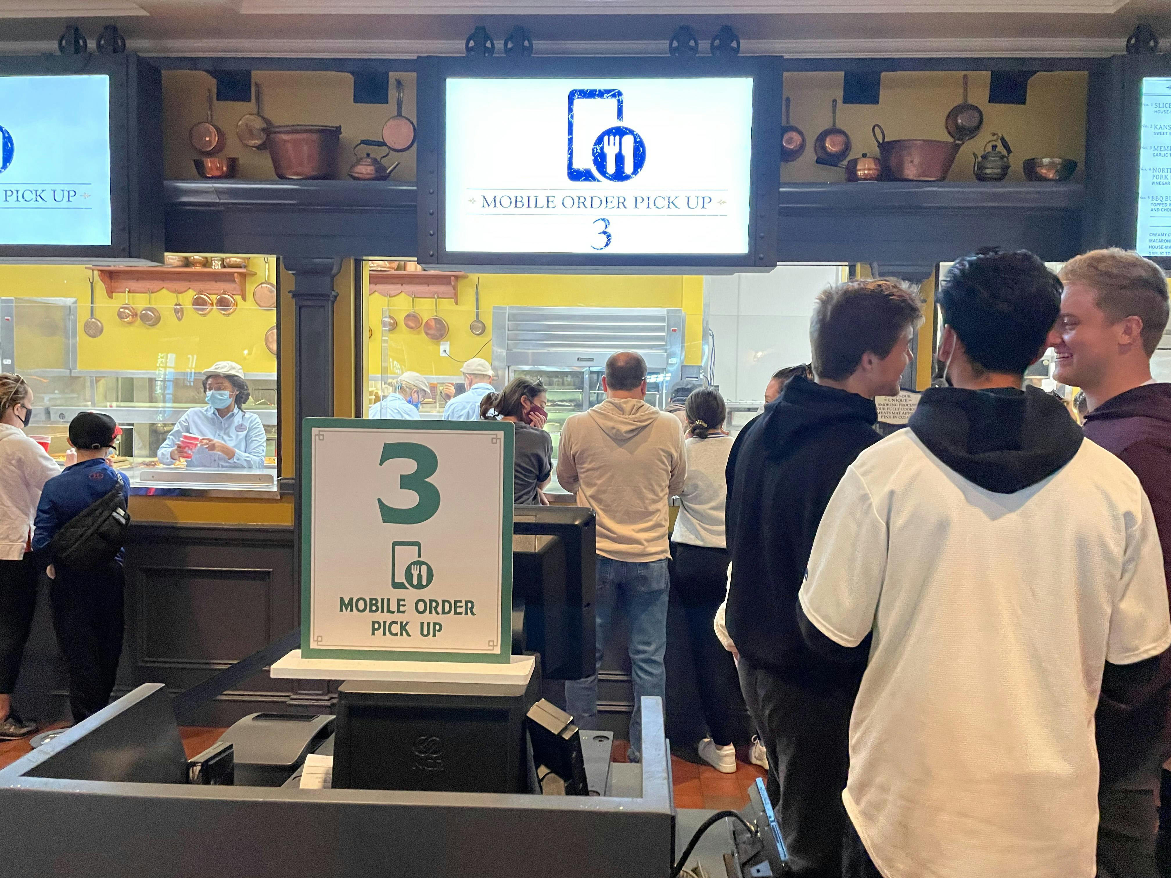 People waiting in line for food at a Disney restaurant with a monitor showing that a mobile order is ready for pick up.