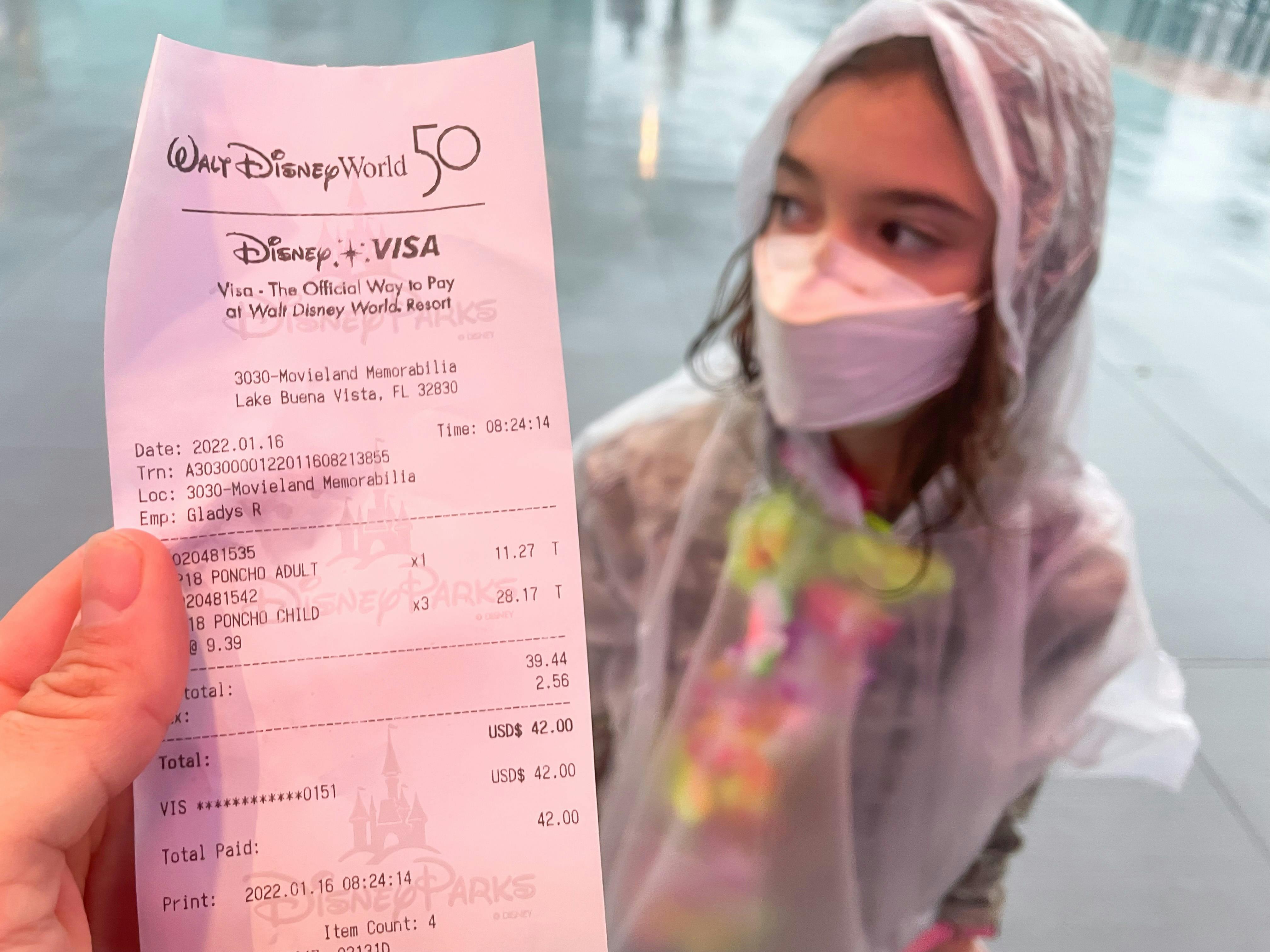 A person holding up a receipt for expensive ponchos from the Disney World park store in front of a child wearing a rain poncho.
