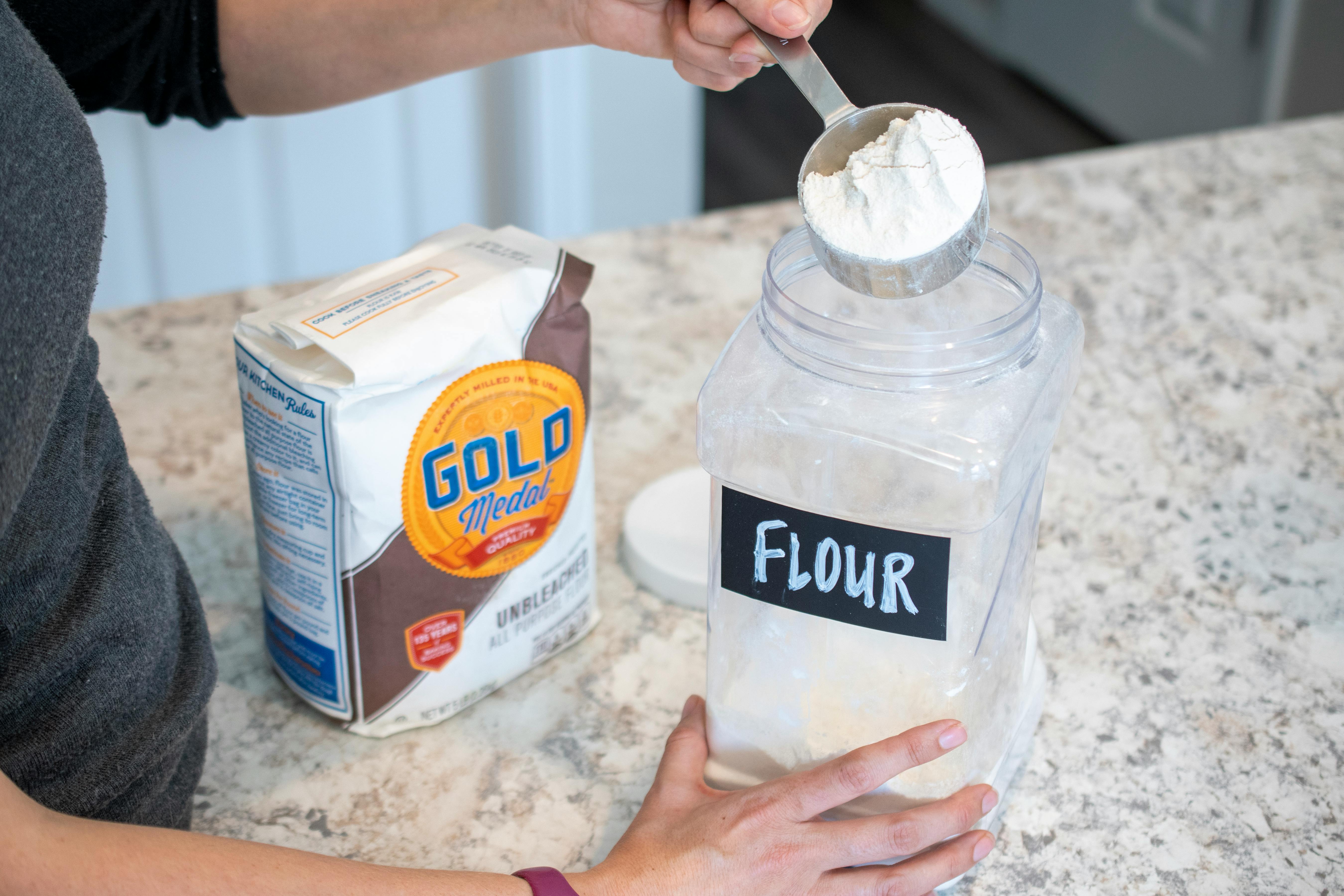 A person scooping flour out of a clear plastic container