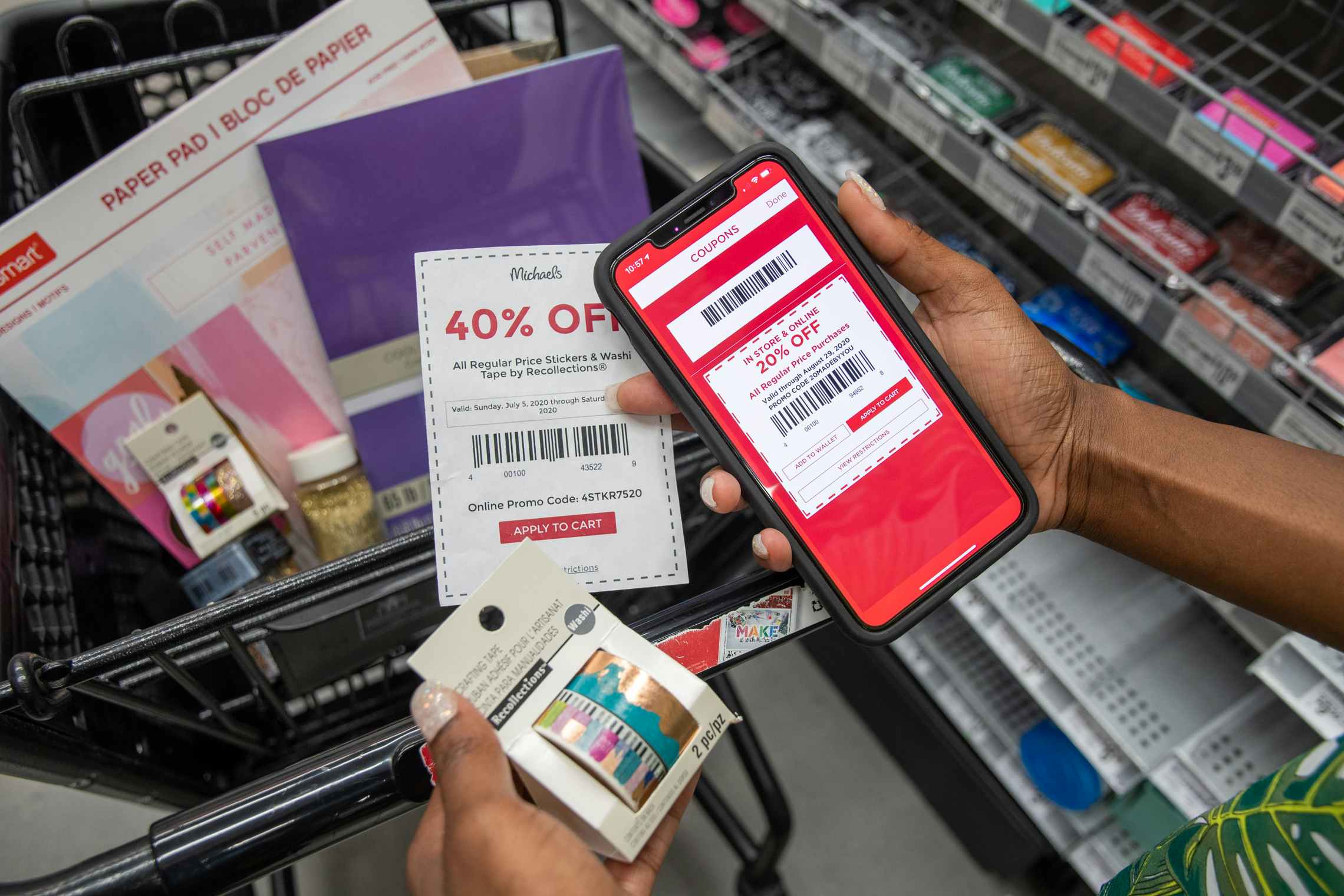 A person holding washi tape, a paper coupon, and an iphone with a second coupon next to a cart filled with products at Michaels.