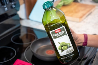 A woman holding a bottle of olive oil next to a stove and frying pan