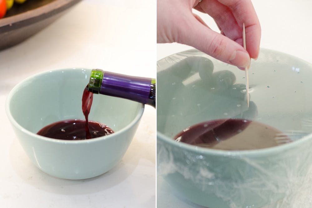 A person pouring wine into a bowl next to a person poking holes into saran wrap covering a bowl of wine.
