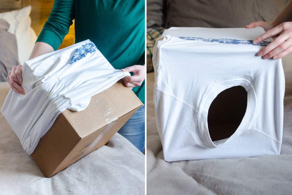 A person putting a t-shirt over a cardboard box, and the cardboard box positioned with the t-shirt head hole as an entrance to the cat hide.