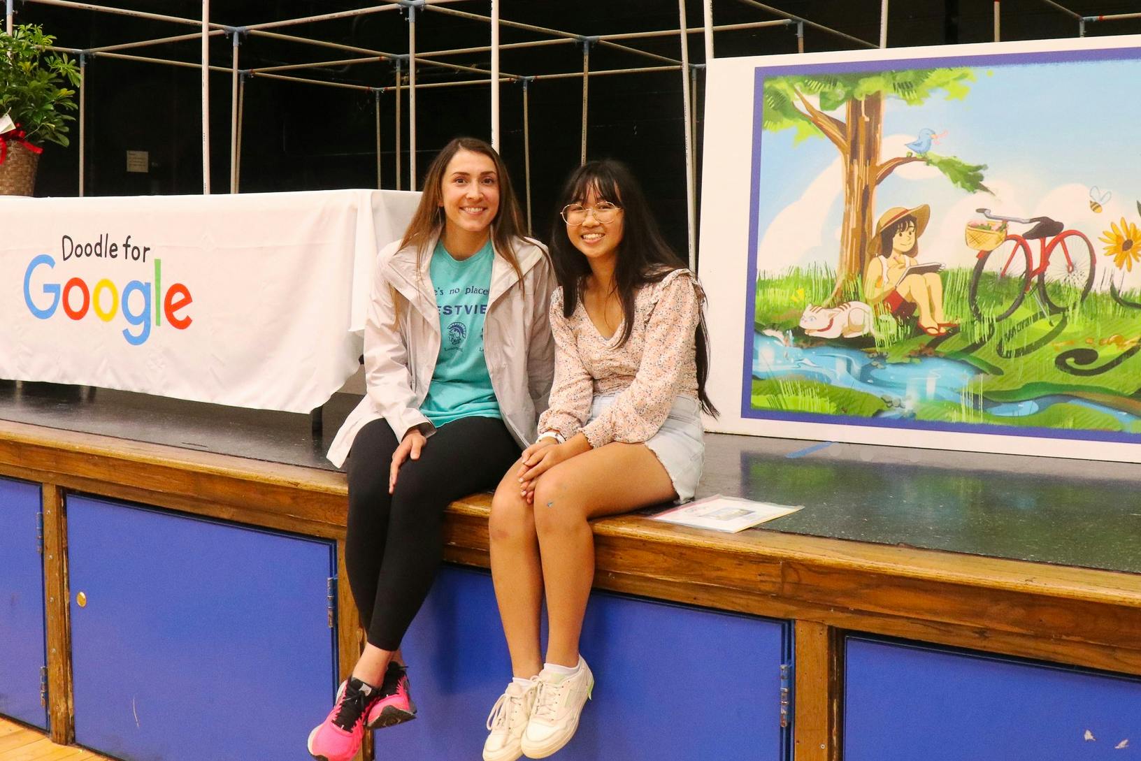 Two students sitting on a stage in front of a student's winning art piece for the state finals of the Doodle for Google scholarship competition.