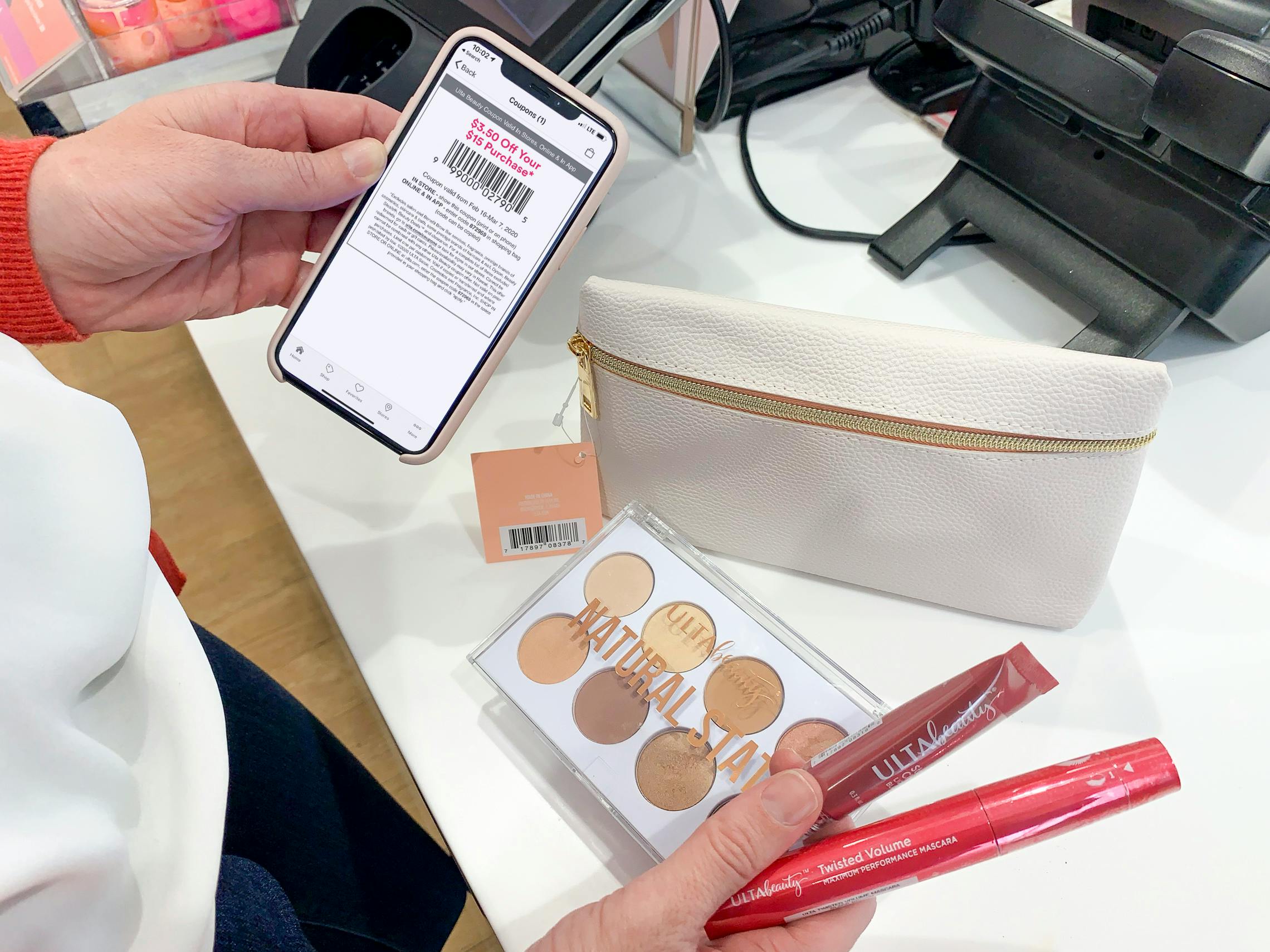 A person holding a cell phone displaying an Ulta coupon at checkout with a free gift and Ulta Beauty Cosmetics on the counter.