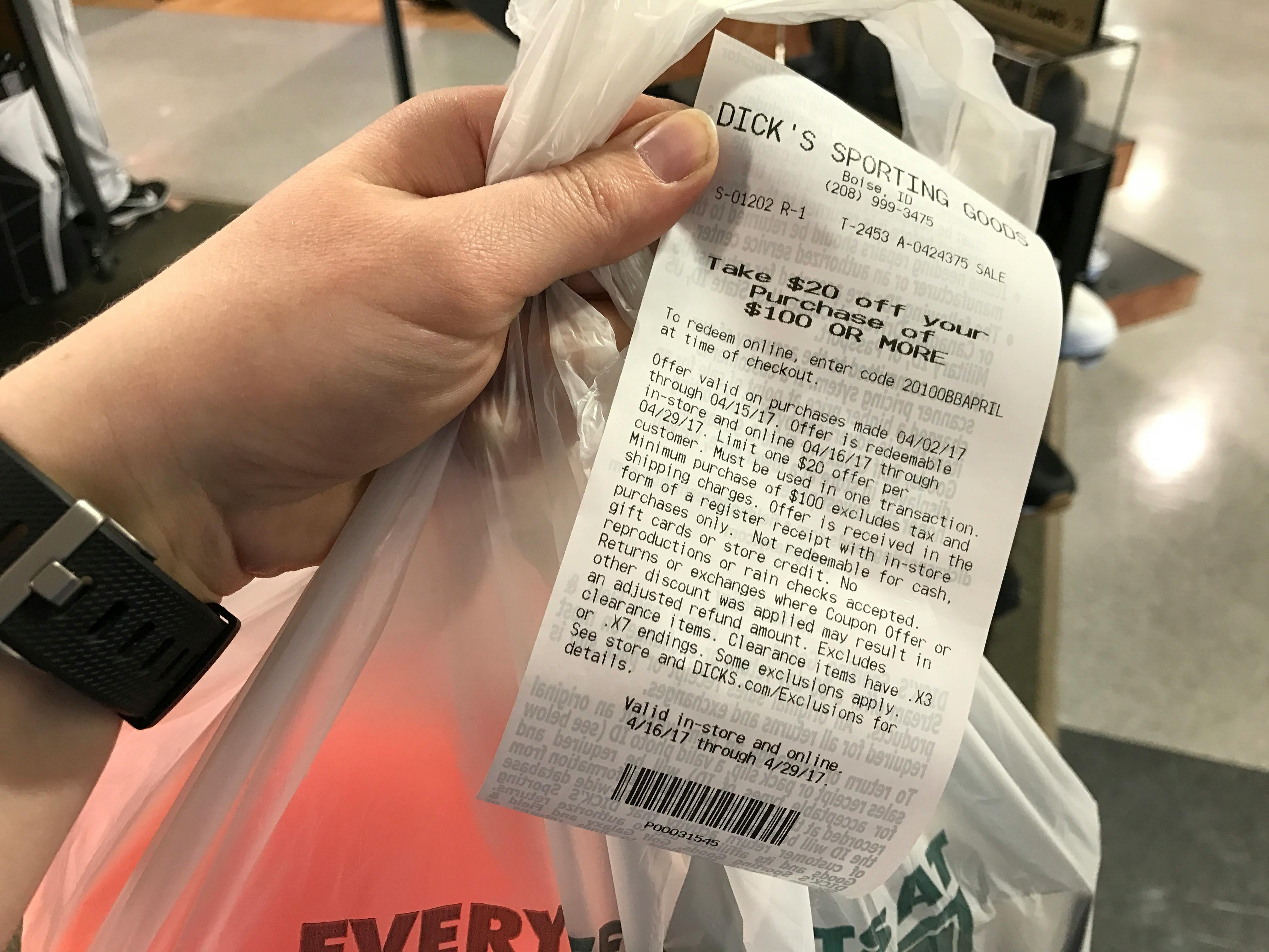 A person's hand holding a Dick's Sporting Goods bag and a receipt with a coupon for $20 off of a purchase of $100 or more.
