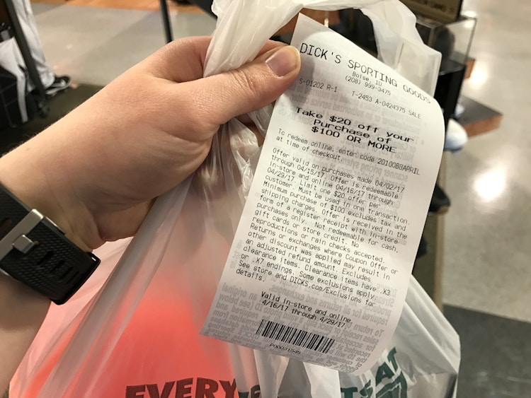 A person's hand holding a Dick's Sporting Goods bag and a receipt with a coupon for $20 off of a purchase of $100 or more.