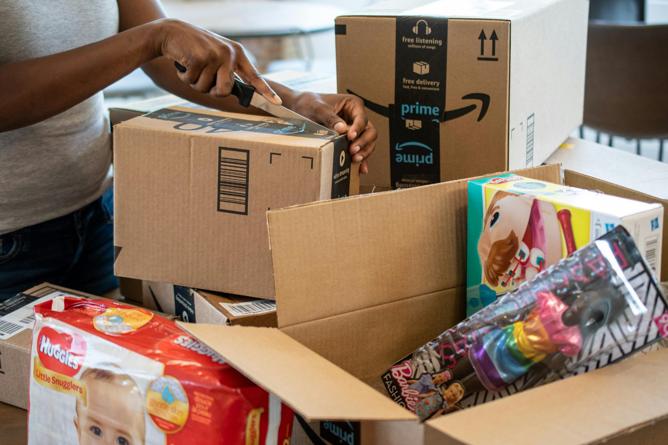 A woman opening an Amazon box with a knife, surrounded by more Amazon boxes with products.