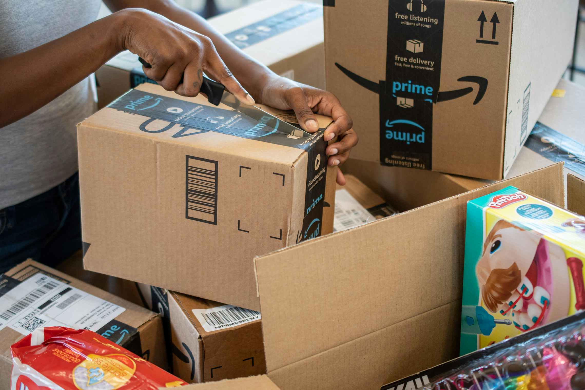 A woman's hands opening an Amazon box with a knife, with more Amazon boxes on the table.