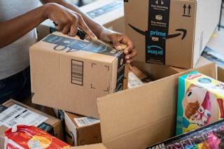 A woman's hands opening an Amazon box with a knife, with more Amazon boxes on the table.