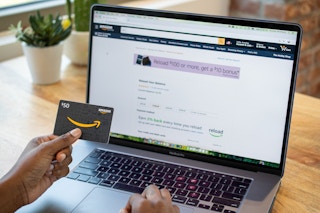 A woman holding an Amazon gift card in front of a computer screen with the Amazon gift card reload page up on it.