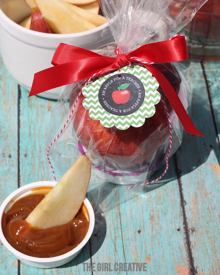 An apple in a clear plastic bag with a ribbon tied around it with a tag that reads an apple for the teacher. in front of a container of caramel with an apple slice sticking out.