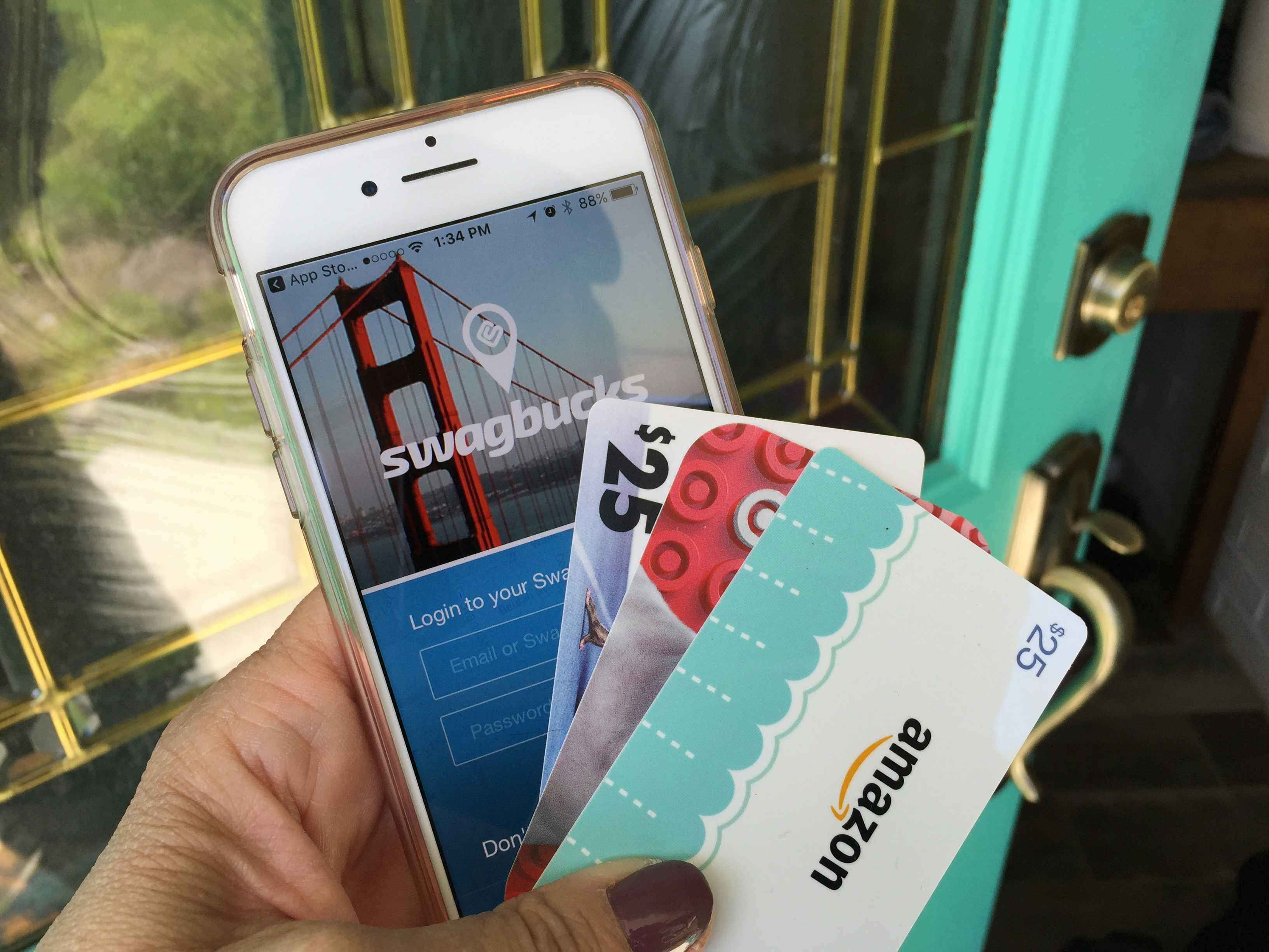 A person's hand holding an iPhone displaying the Swagbucks mobile app, a gift card for $25, a target gift card, and an Amazon gift card for $25, in front of an open door.