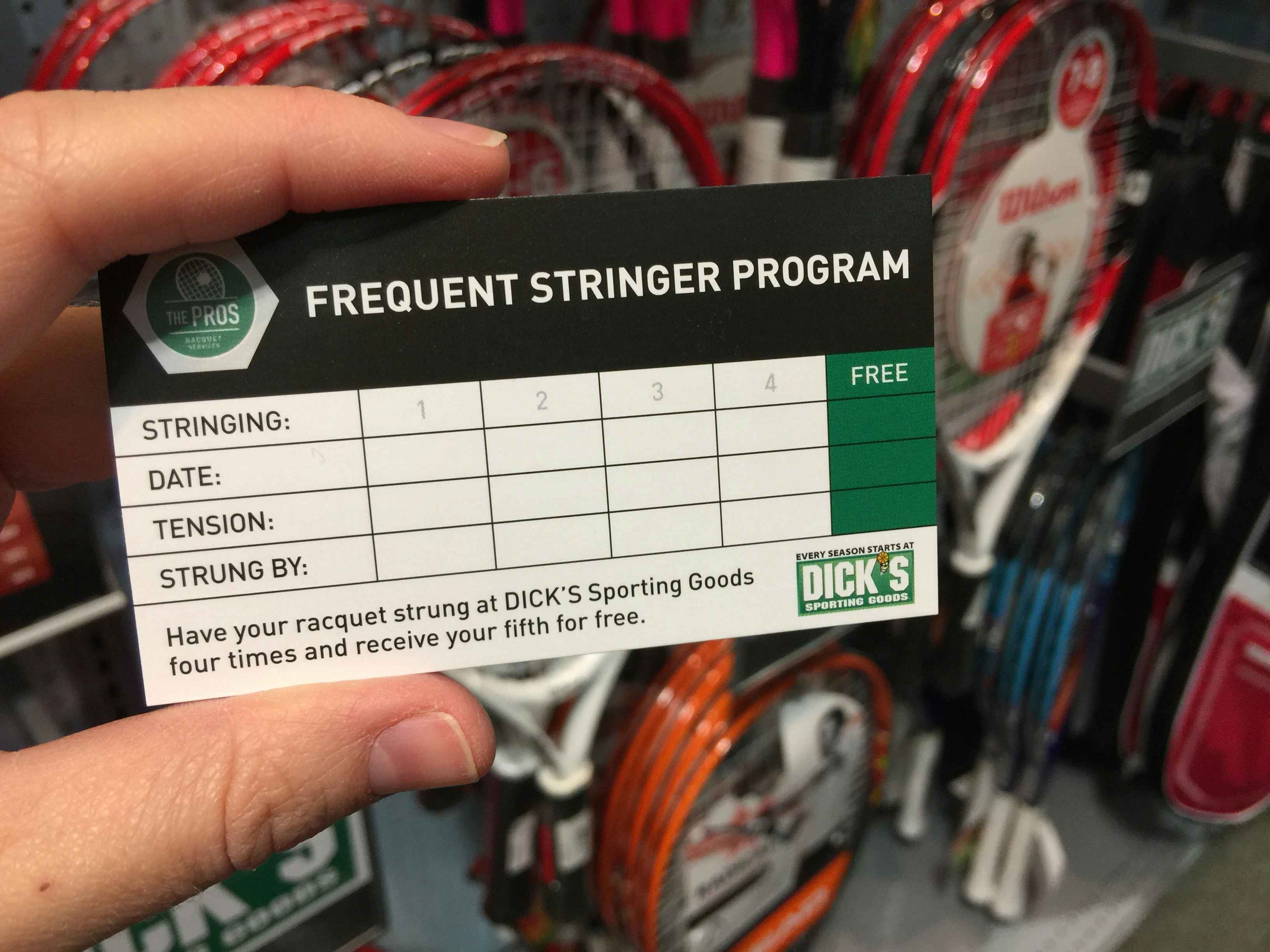 A person's hand holding up a Frequent Stringer Program card in front of a wall of rackets inside Dick's Sporting Goods.